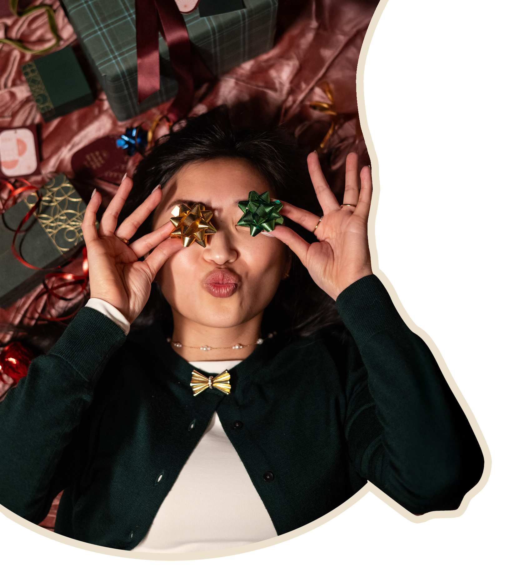 A woman lying down surrounded by wrapped Christmas presents, holding two gift bows over her eyes, one gold and one green, with her lips puckered.