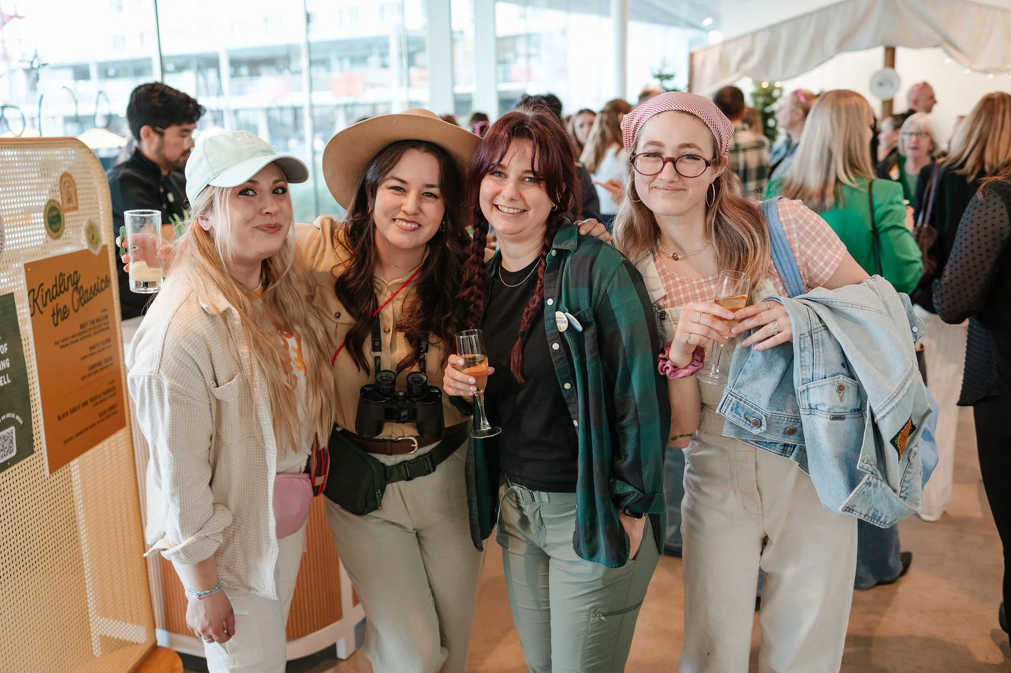 Four women standing together at an indoor event, smiling and holding drinks, with a crowd in the background.