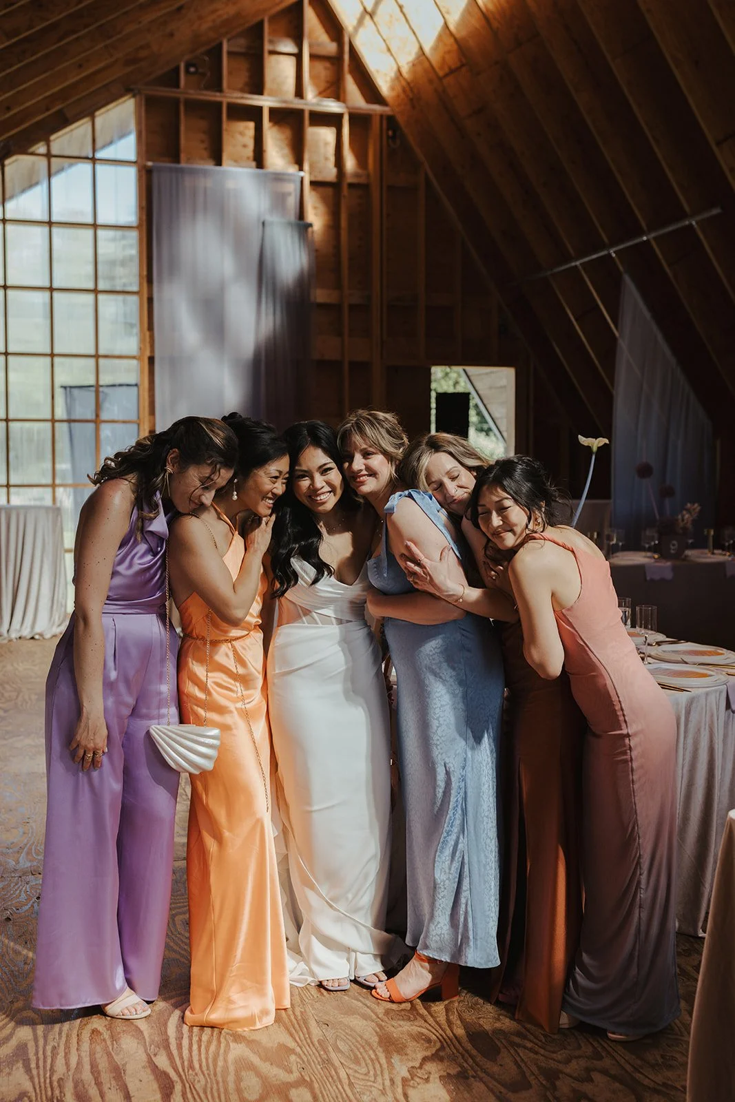 Group of women hugging and smiling at a celebration or wedding reception inside a wooden venue with large windows.