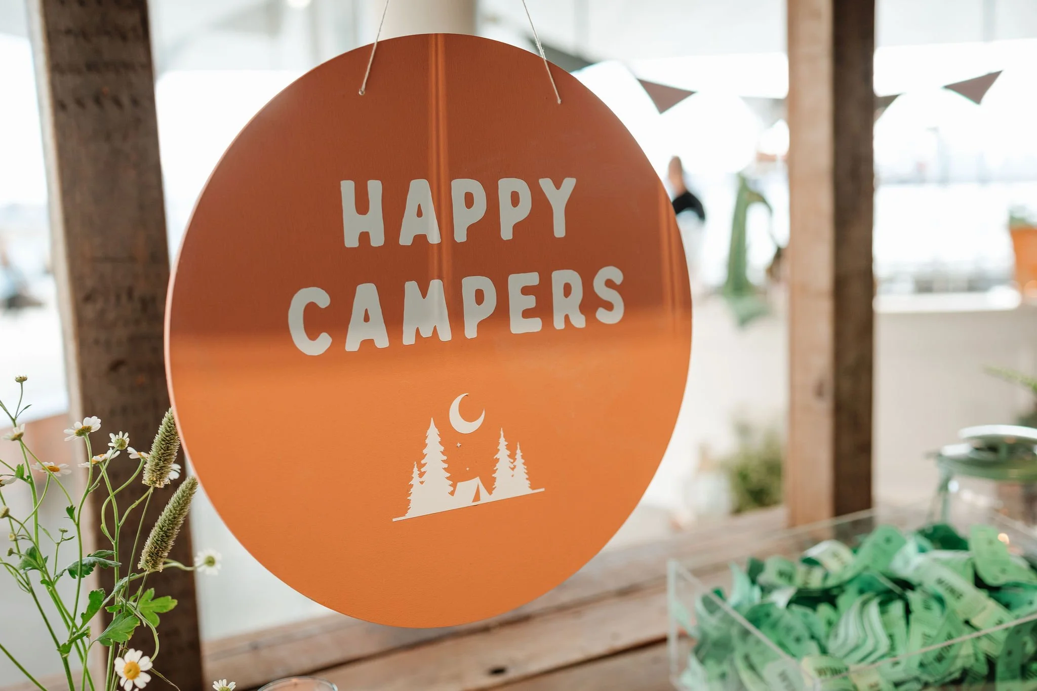 Orange circular sign with white text saying 'Happy Campers' and a graphic of a tent, trees, and a crescent moon, hanging from a string indoors.