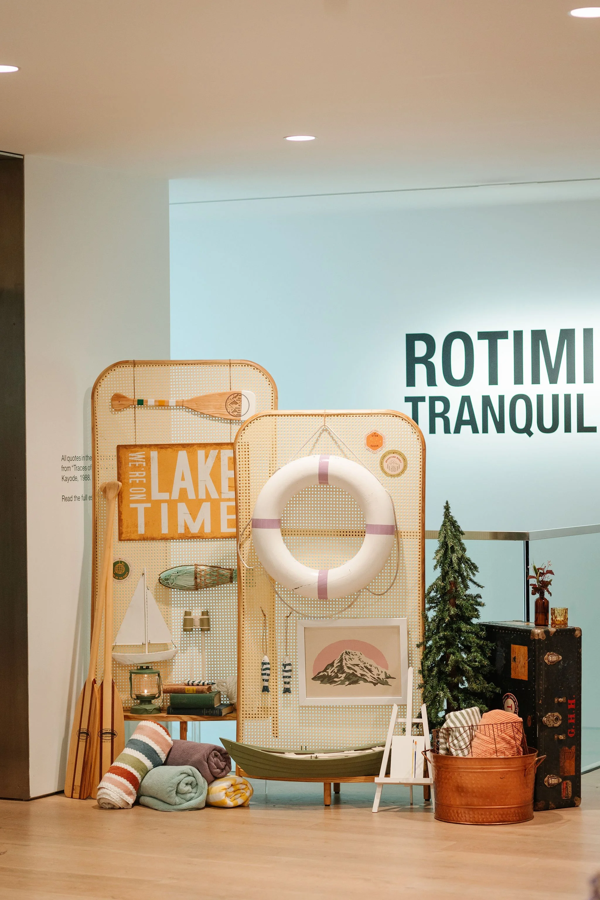 Decorative display with boating and outdoor themed items, including a life ring, a small boat, rolled towels, picnic basket, miniature tree, and nautical accessories, set against a wall with text.