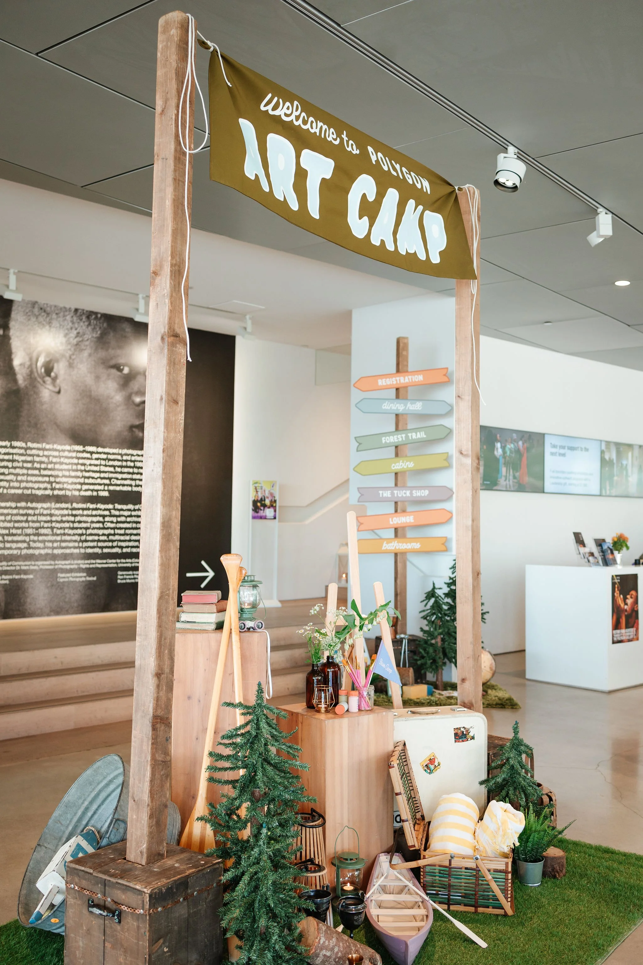 Decorative welcome sign for Art Camp with a wooden frame, surrounded by small trees, lanterns, and camping-themed items, inside an art gallery or community center.