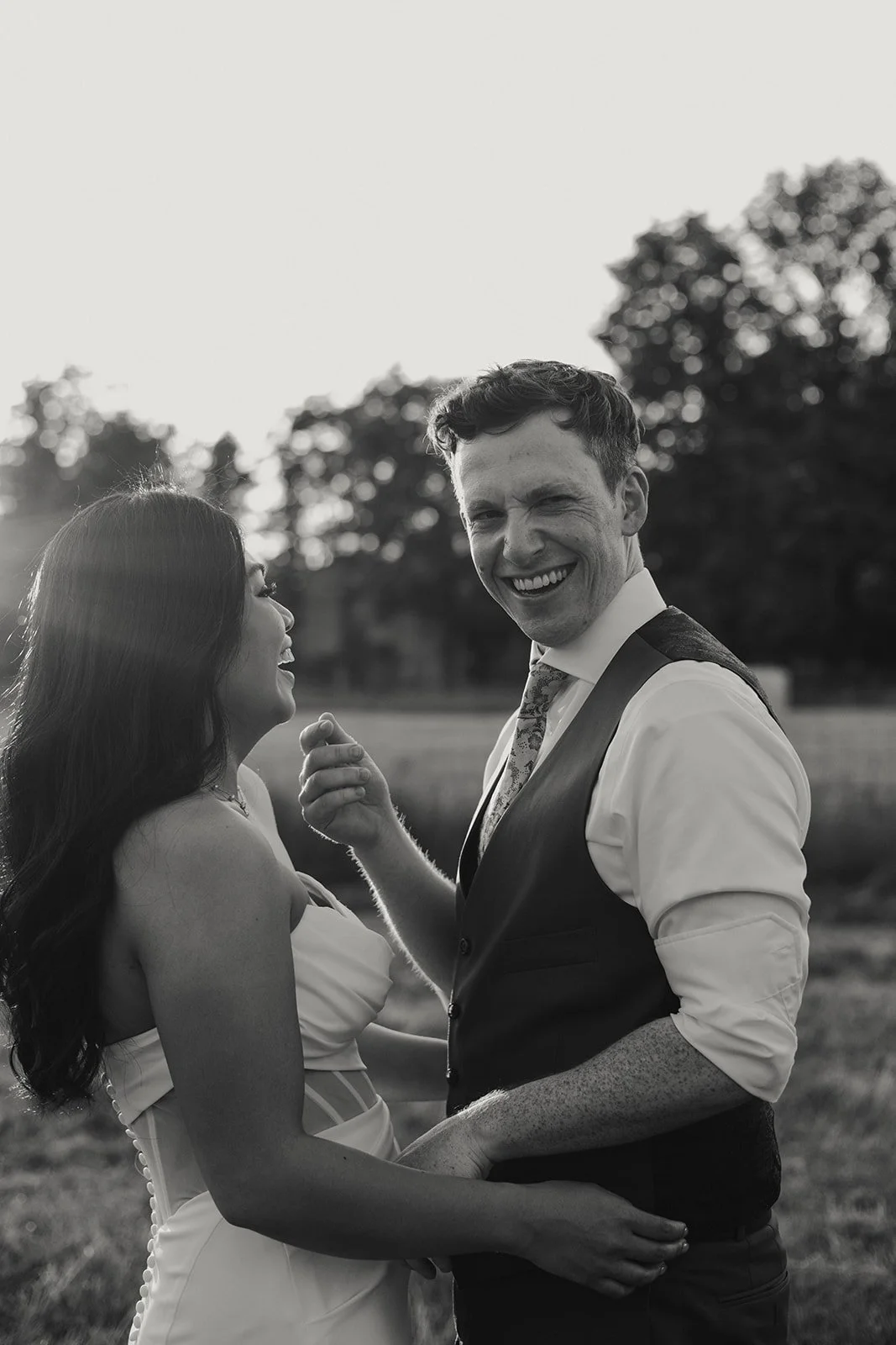 A black-and-white photo of a smiling man and woman outdoors, standing close, with the man holding the woman’s waist and the woman looking at the man, backlit by the sunset and with trees in the background.