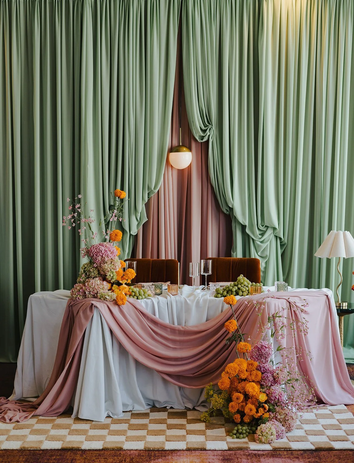 Elegant banquet table with pink and white draped fabric, floral arrangements, grapes, and glassware, set against a backdrop of green curtains with a pink curtain in the middle.