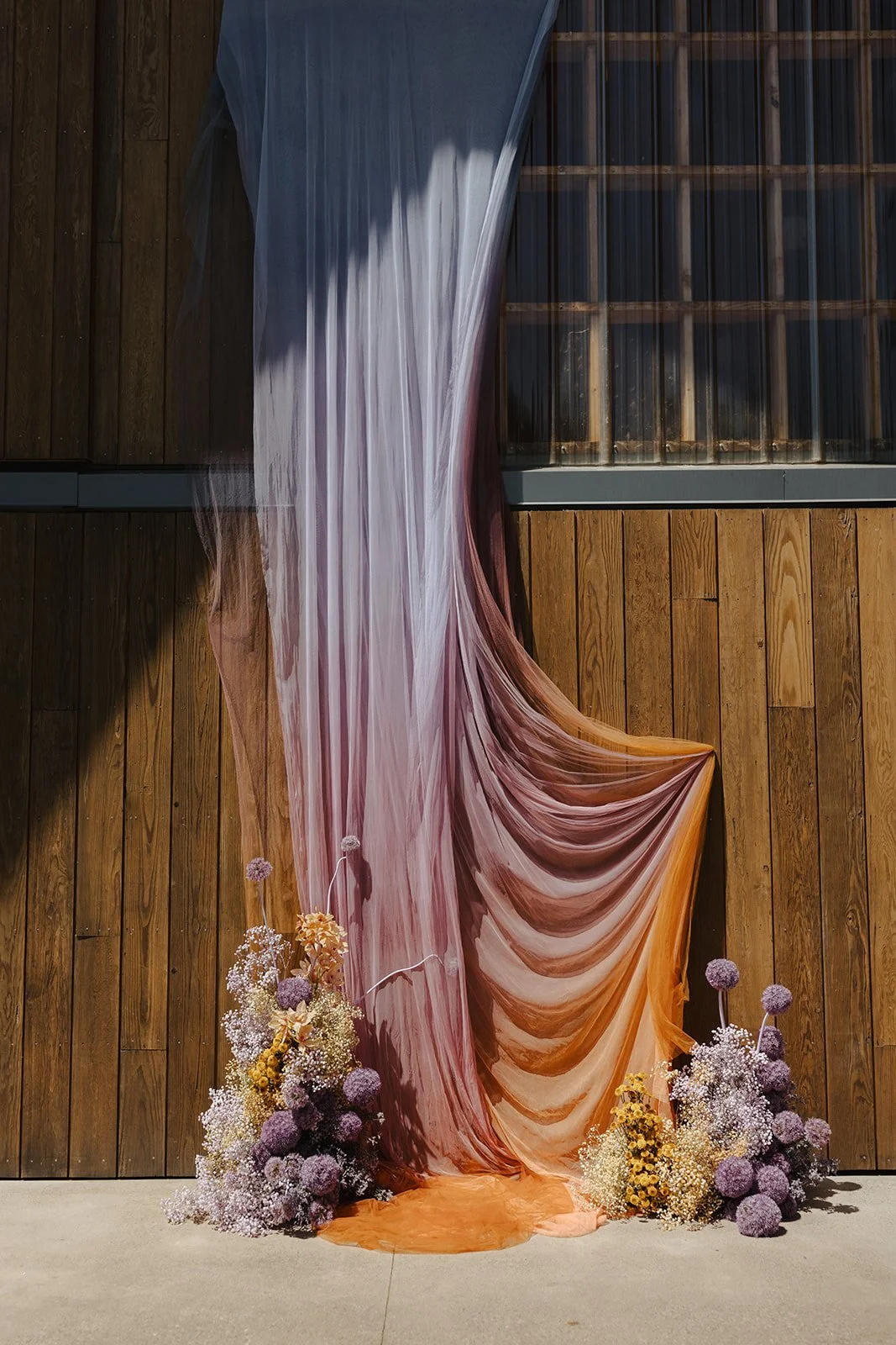 Decorative fabric drapes in shades of purple, pink, and orange hanging against a wooden wall, adorned with bouquets of purple and white flowers at the base.