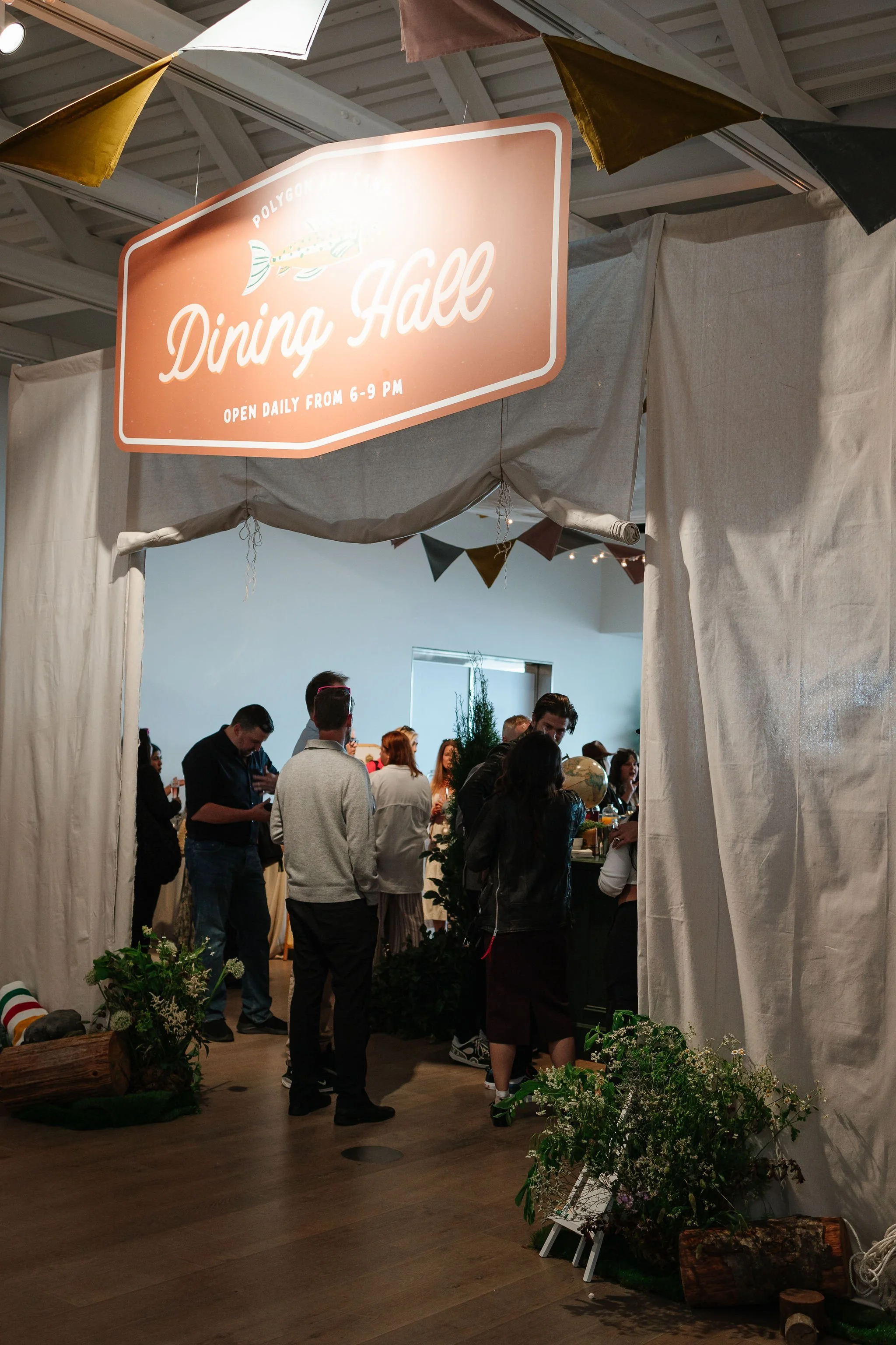 People gathered inside a dining hall with a sign above them reading 'Dining Hall' and a fish illustration, open daily from 6 to 9 pm, decorated with a nautical theme and foliage.