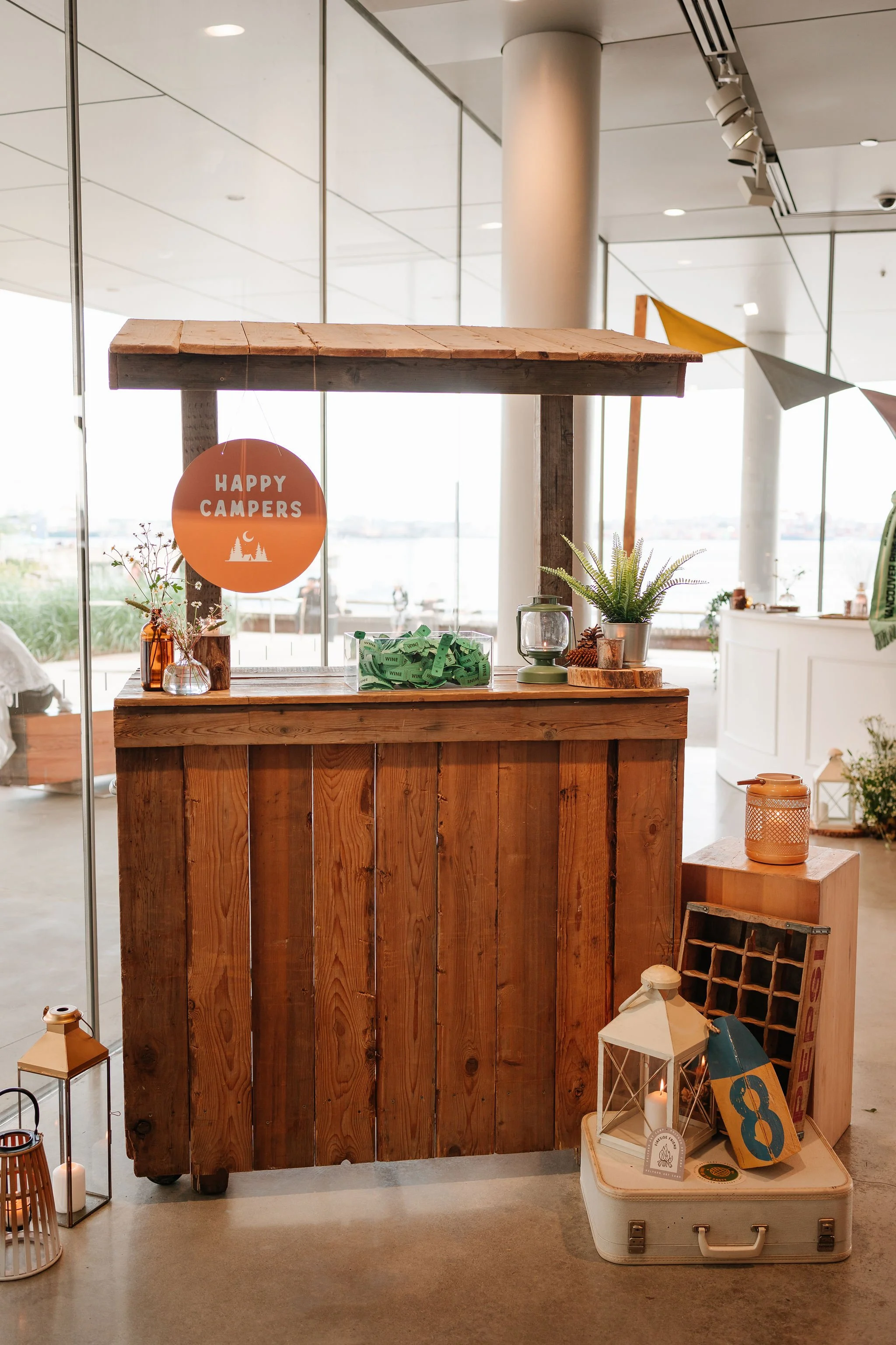 Wooden table with a sign that reads 'Happy Campers', decorative candles, plants, and a container of small green ribbons inside a brightly lit modern indoor space with large windows.