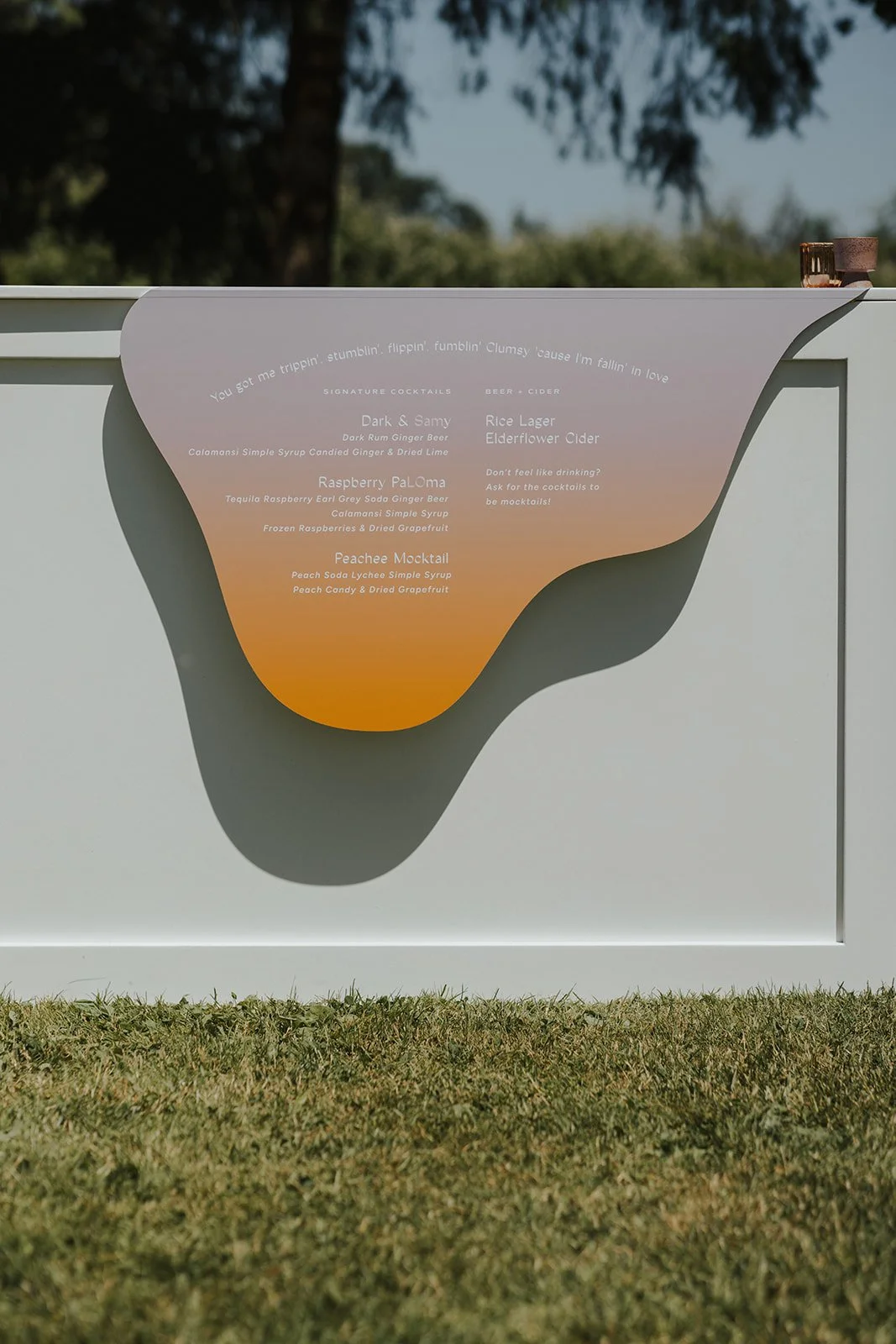 A menu displayed on a white board outdoors with a sunset background, featuring cocktails and drinks like 'Dark & Samy,' 'Rice Lager Elderflower Cider,' 'Raspberry PaLoma,' and 'Peach Mocktail,' with colorful gradient design mimicking a sunset.