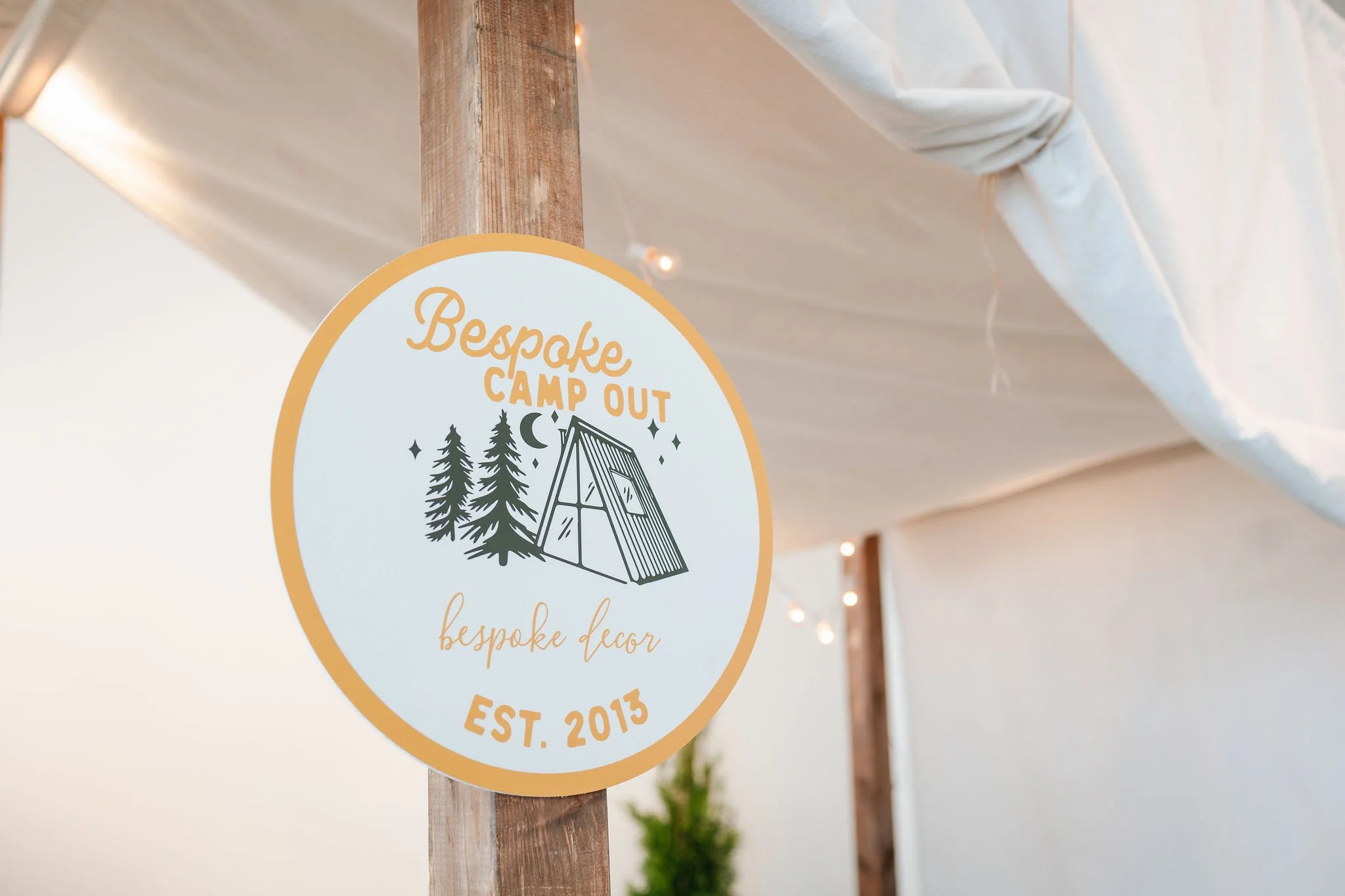A circular sign with the text 'Bespoke Camp Out', 'bespoke decor', 'EST. 2013', hanging from a wooden post inside a tent with string lights.