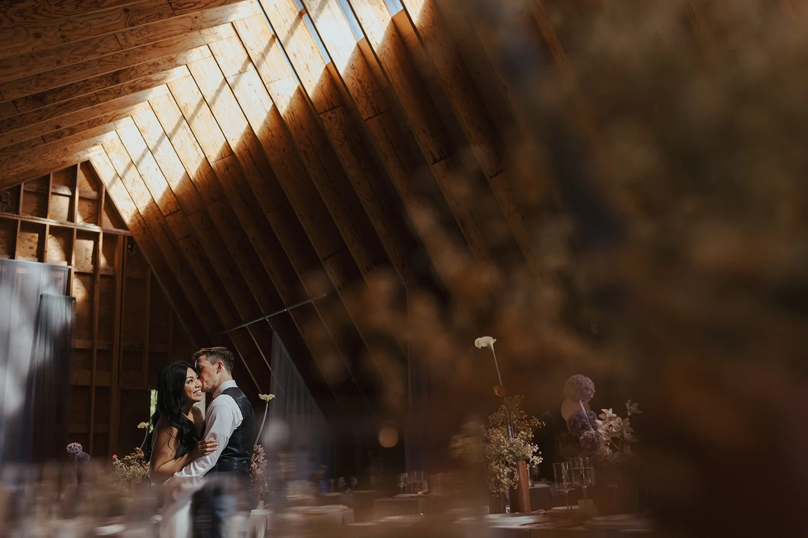 A couple shares a quiet moment at their wedding reception in a wooden-paneled venue, surrounded by floral arrangements and elegant table settings.