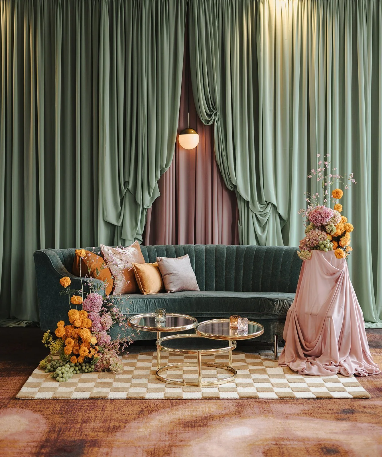 Elegant living room with green curtains, a dark green velvet sofa with pastel cushions, floral arrangements, and round glass coffee tables on a checkered rug.
