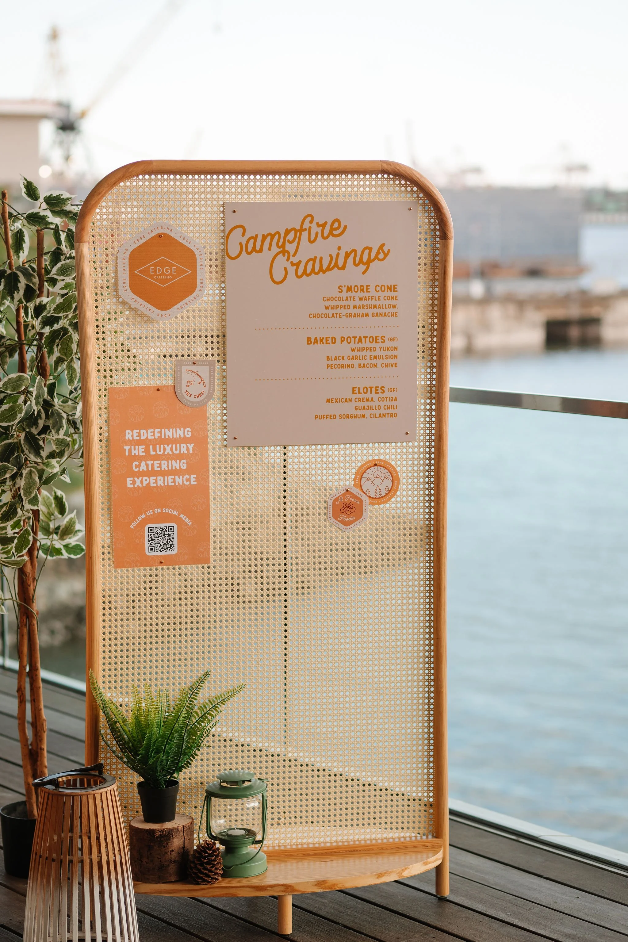 A campfire catering menu board listing s'more cone, baked potatoes, and elotes, with decorative plants and a lantern on a wooden table near a body of water.