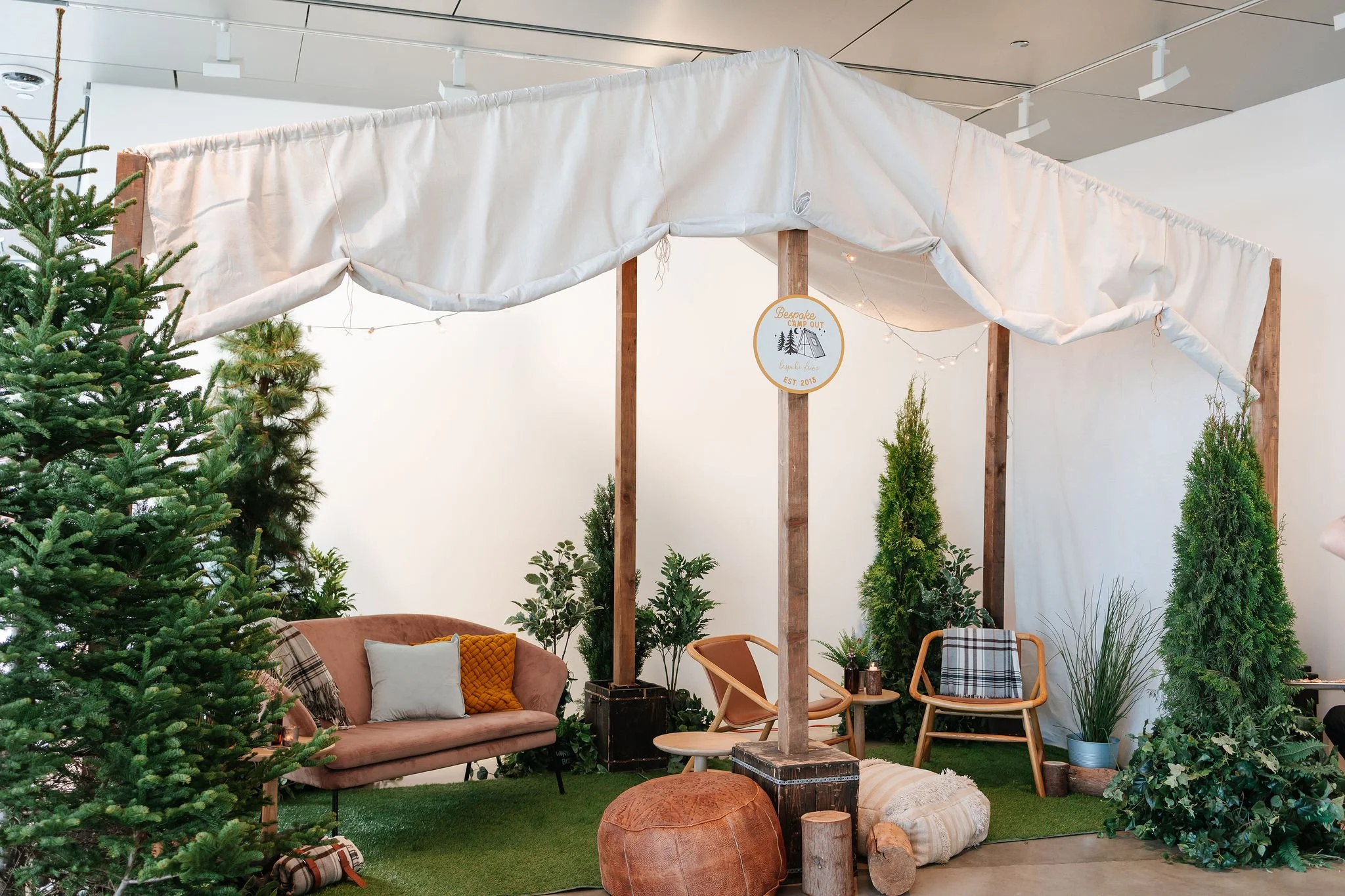 Indoor display with a white canopy, a pink sofa with pillows, wooden chairs, potted plants, a brown pouf, and evergreen trees, creating a cozy, nature-inspired setting.