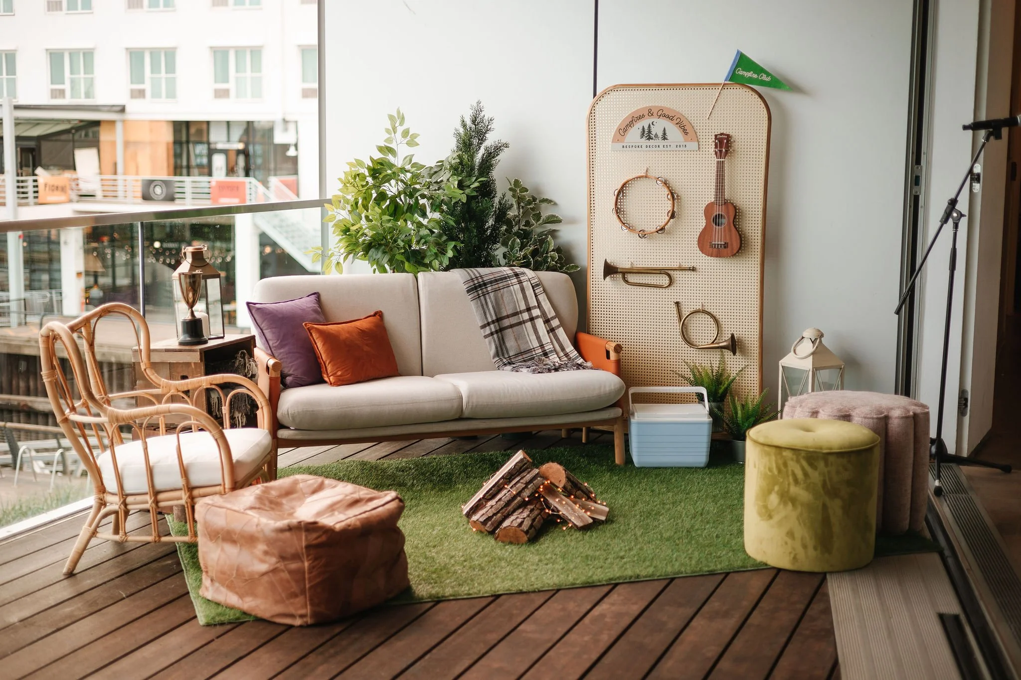 Cozy indoor living space with rattan armchair, beige sofa with purple and orange pillows, green plants, and a pegboard wall with musical instruments and decor. There is also a green ottoman, a pink pouf, a small lamp, and a framed lantern. A fire pit