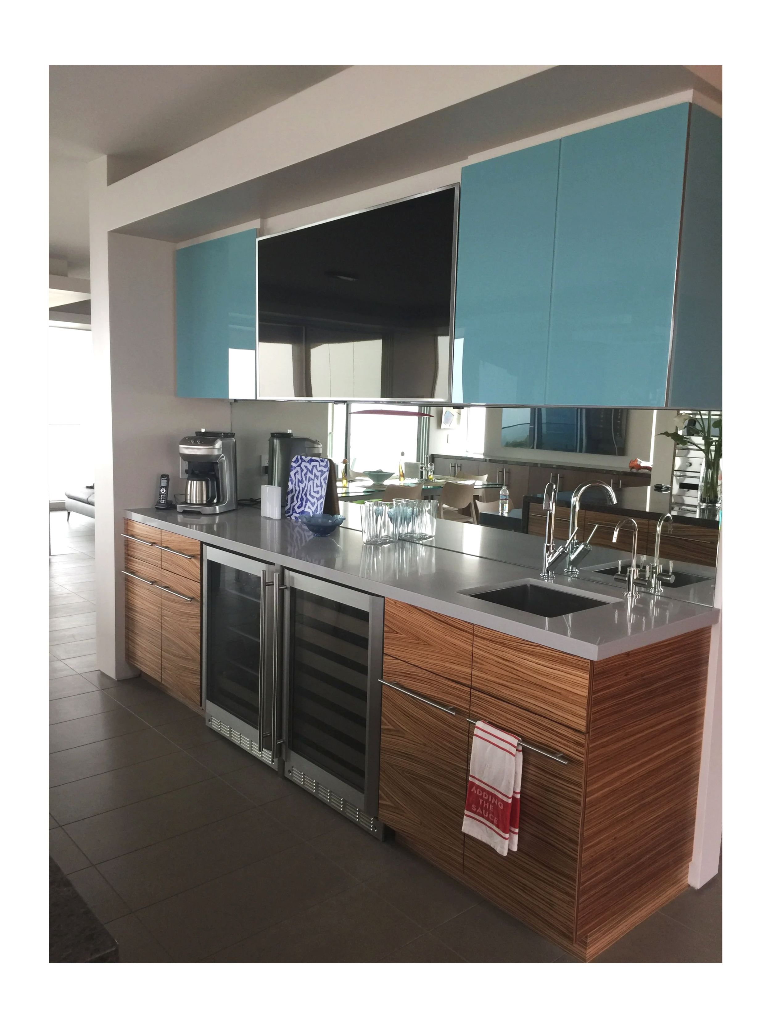 Modern kitchen with wooden cabinets, a white countertop, a small sink, a glass-front wine cooler, and a mirror reflecting a dining area and windows with outdoor views.