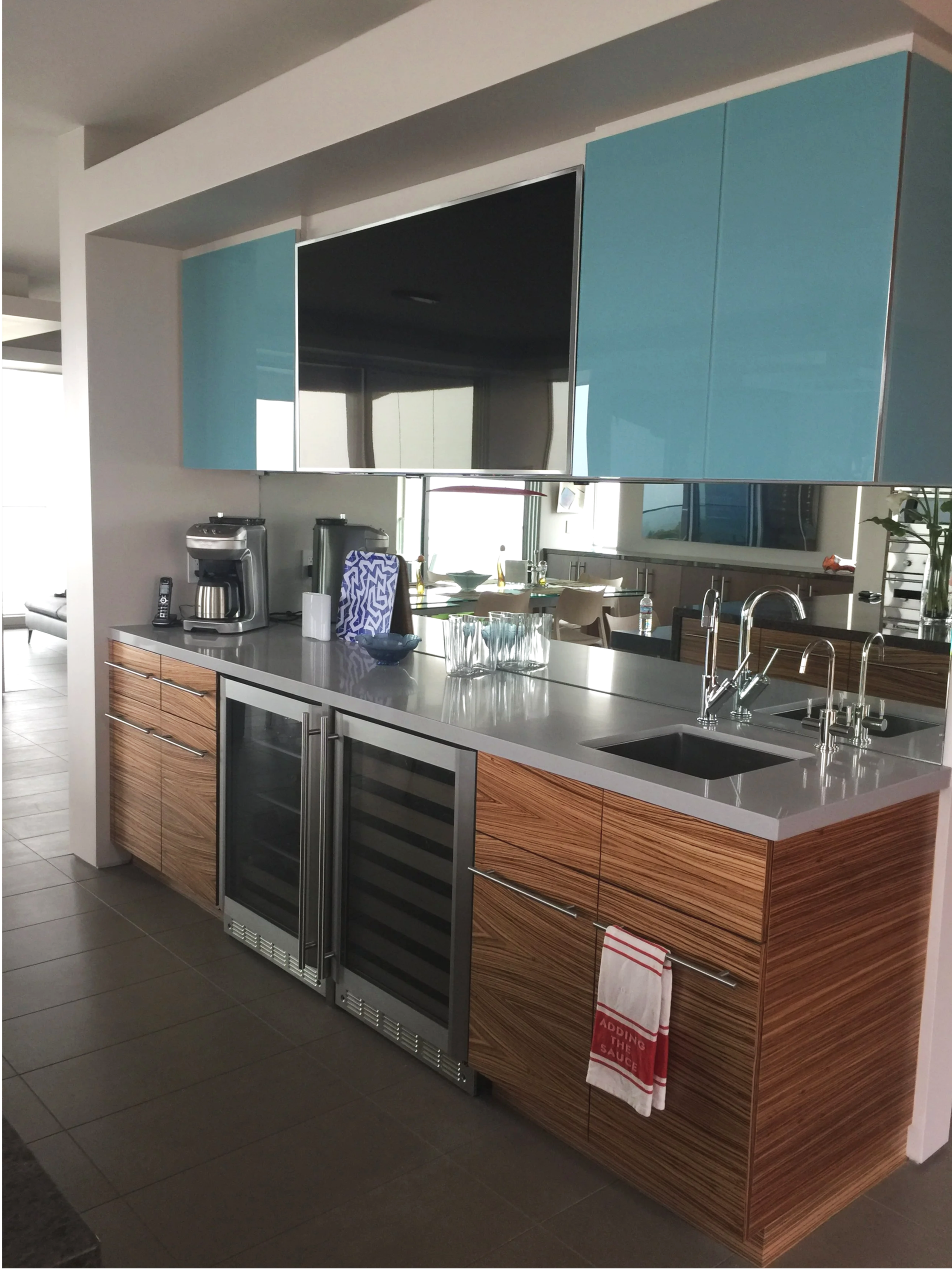 Modern kitchen with wooden cabinets, a white countertop, a small sink, a glass-front wine cooler, and a mirror reflecting a dining area and windows with outdoor views.
