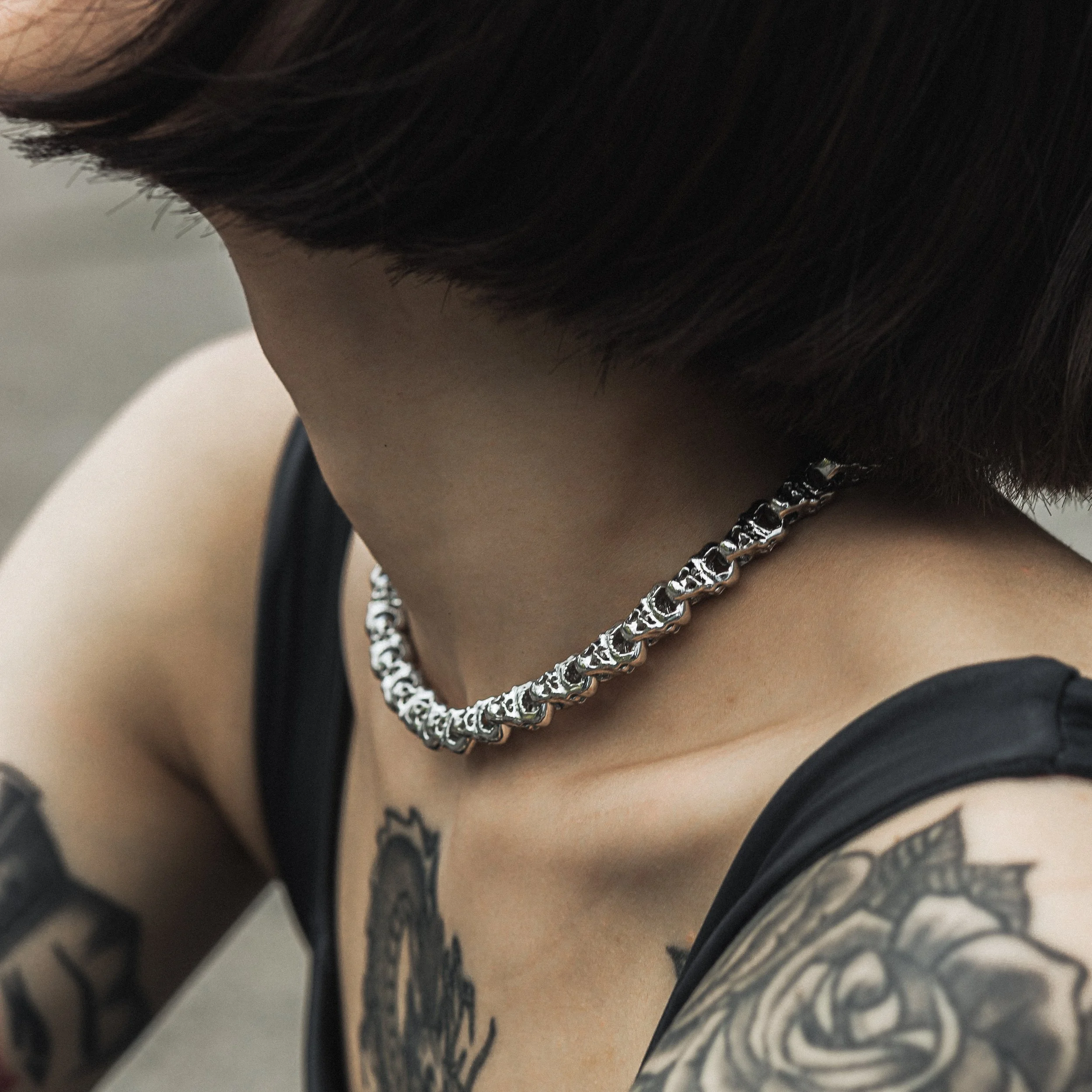 Close-up of a person's neck wearing a silver chain necklace, with short dark hair and tattoos on their shoulder and arm.