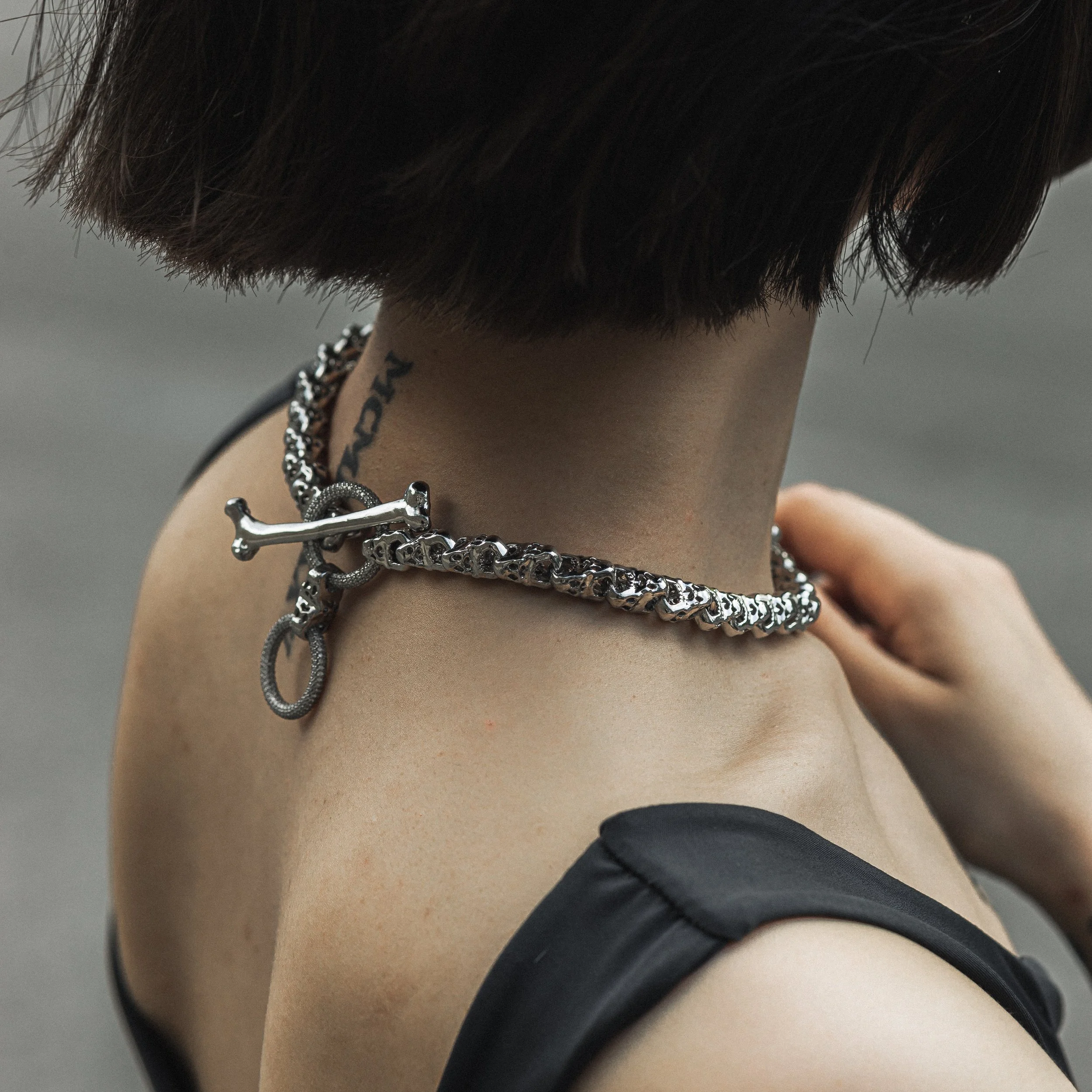 Close-up of a person wearing a chunky silver chain necklace with a dog bone and O-ring charms, seen from behind.
