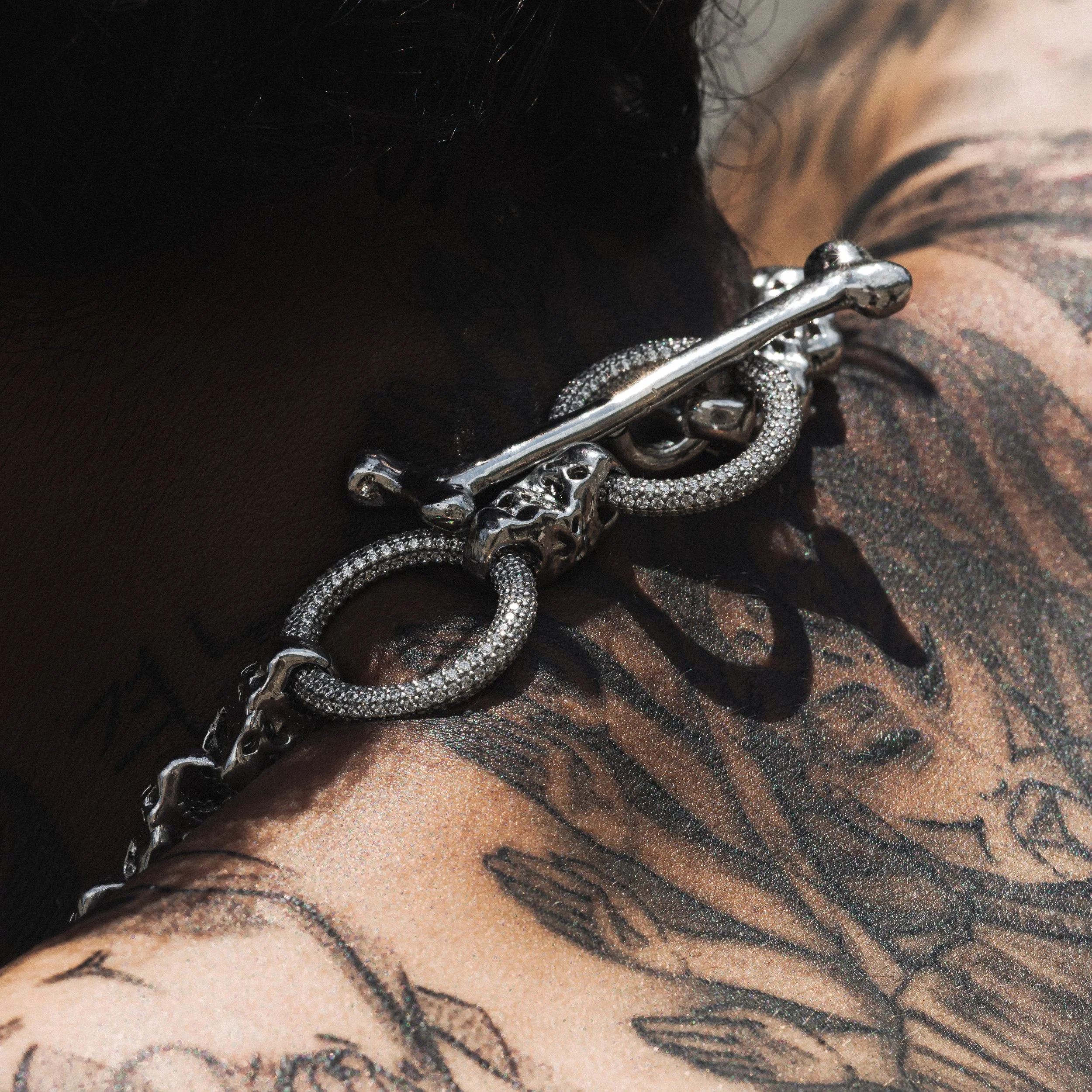 A close-up of a tattooed arm and shoulder with a silver chain and femur bone clasp, featuring intricate designs and dark ink.