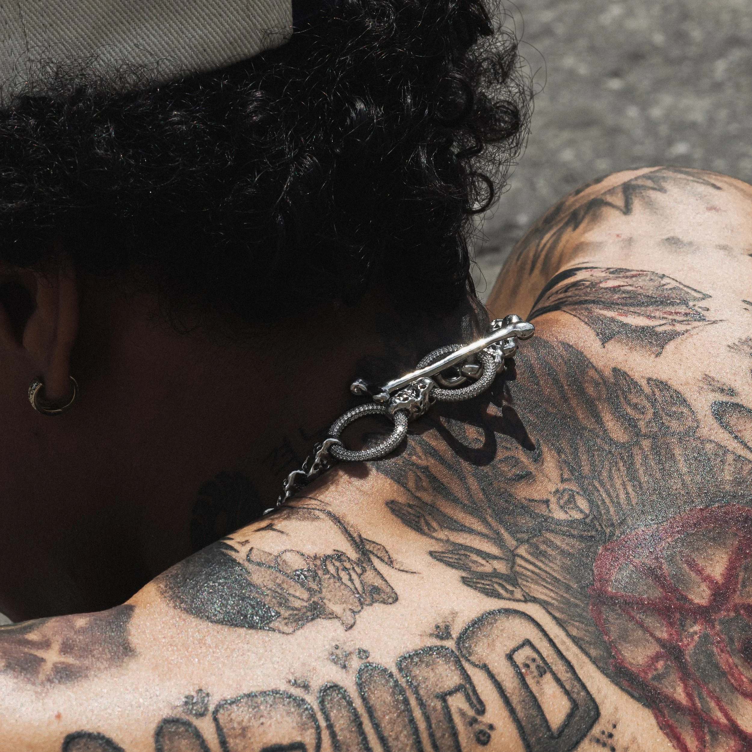 Close-up of a person's shoulder and head, showing many tattoos, a silver chain, and curly black hair.