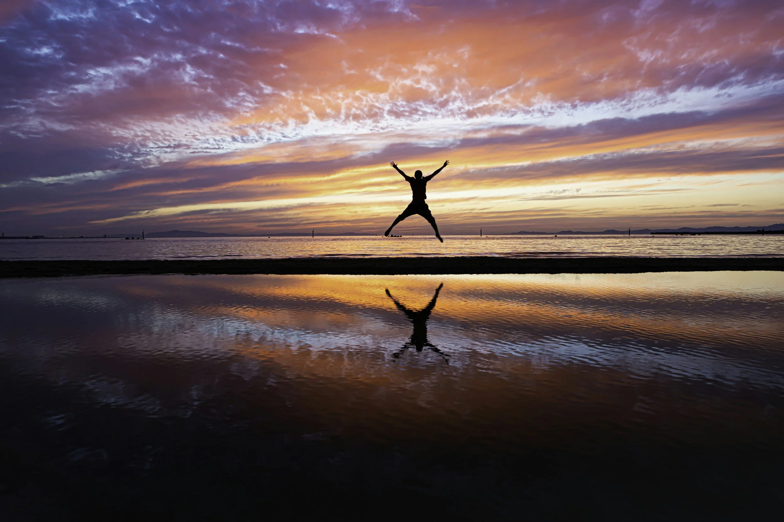 Person jumping on the beach during a colorful sunset with vibrant pink, orange, and purple clouds reflected in the water.