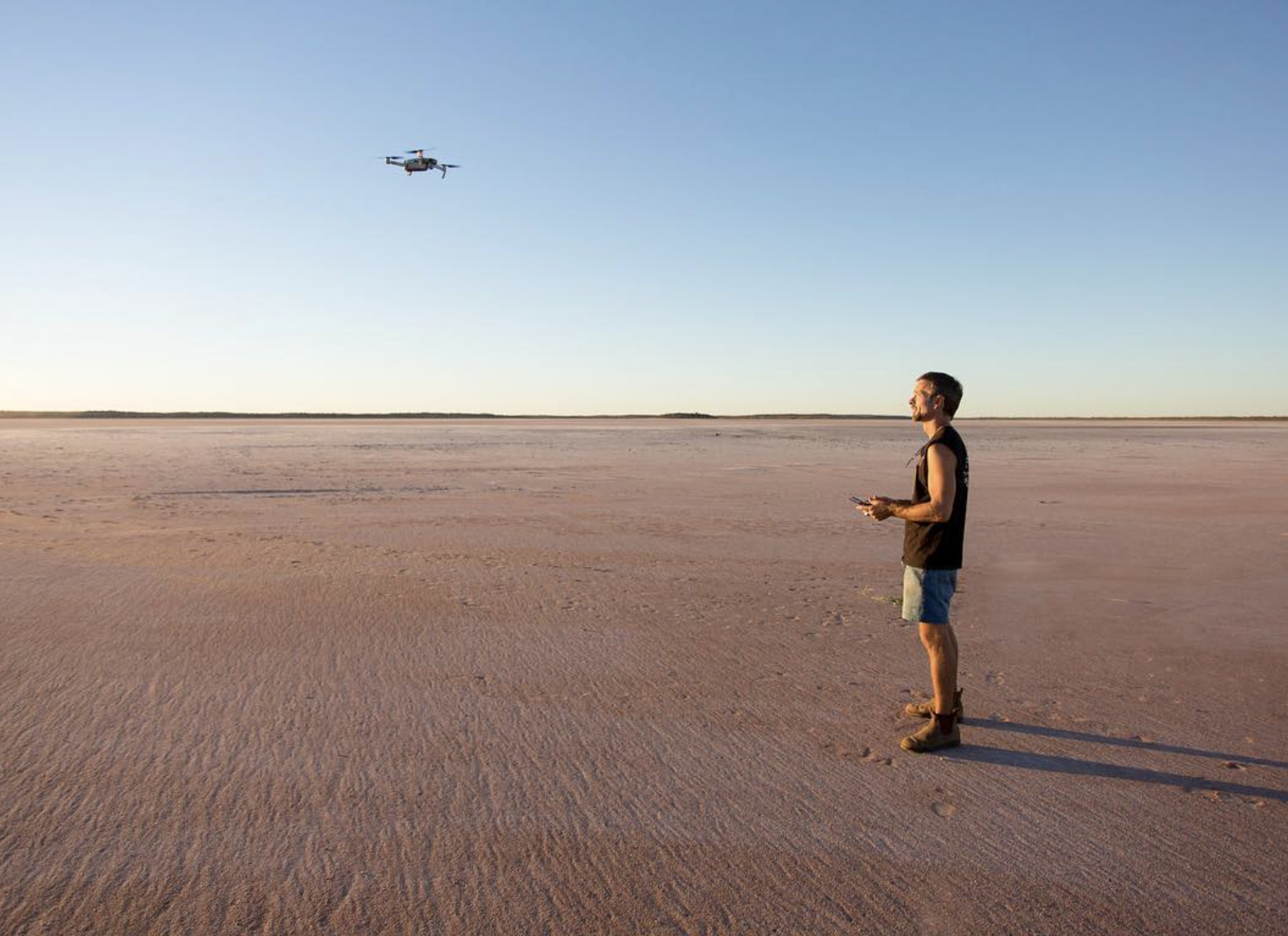 A person standing on a flat, sandy terrain while controlling a drone flying nearby in clear weather.