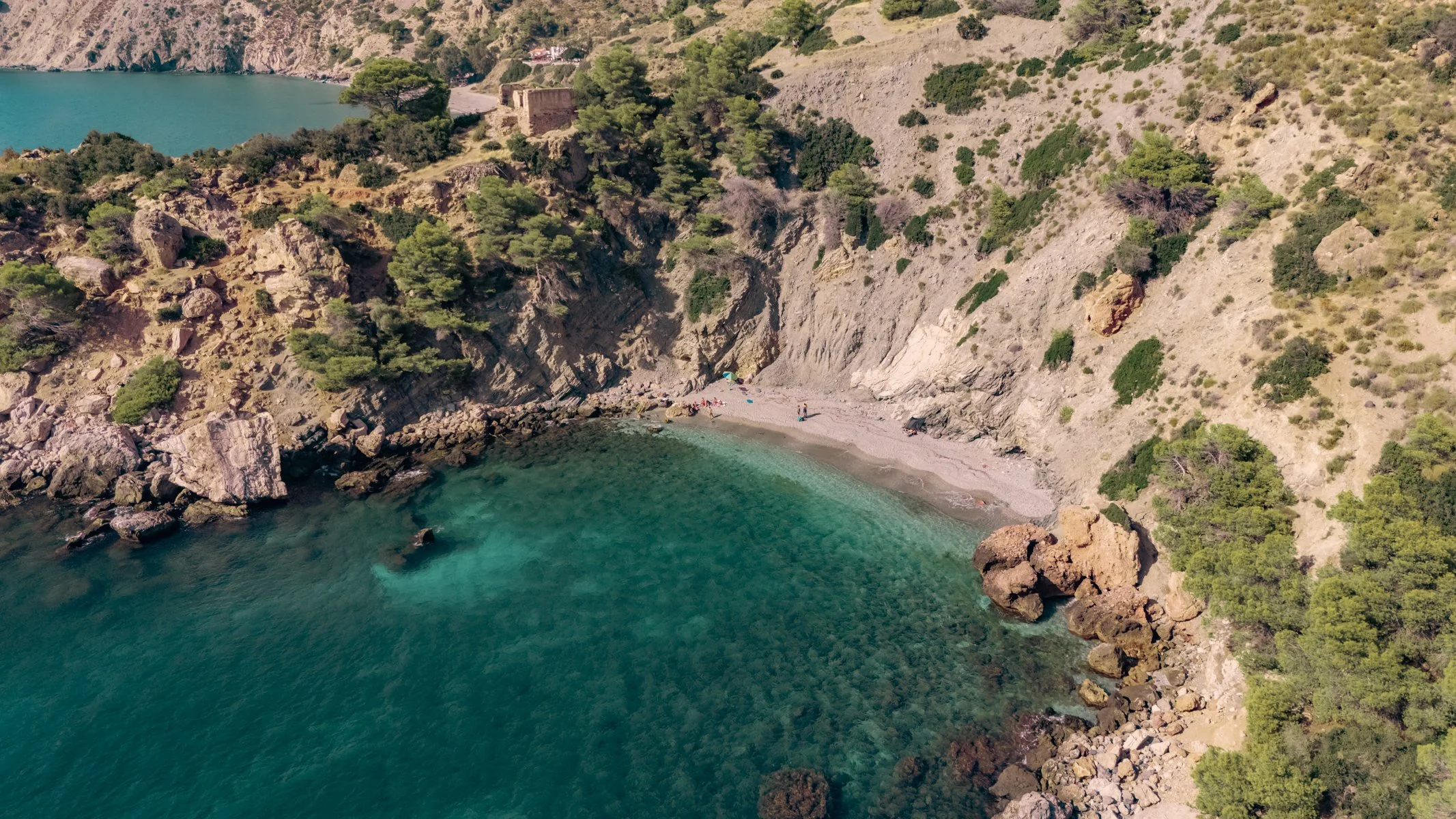 A small secluded beach surrounded by rocky cliffs with green trees, turquoise water, and a few people relaxing under umbrellas.
