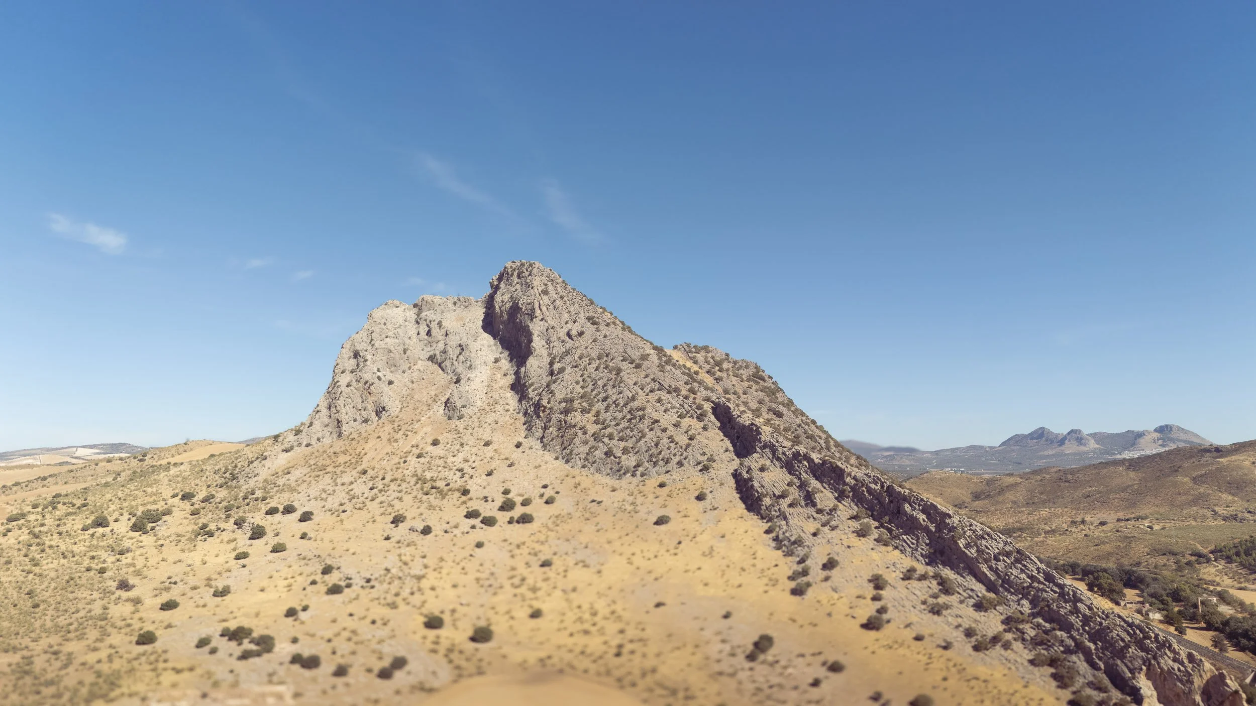 A rugged mountain peak with steep slopes, partially covered in sparse vegetation, under a clear blue sky with a few small clouds.
