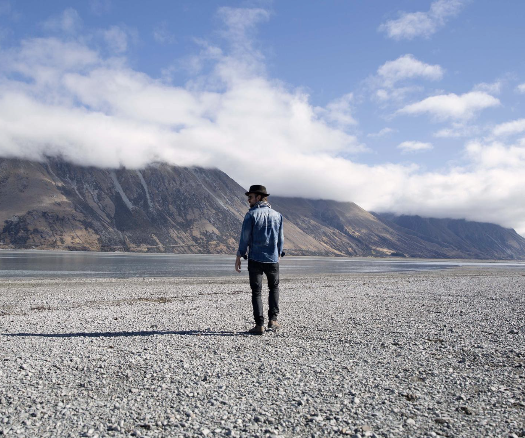 Man in denim jacket and hat walking on rocky beach near a lake with mountains and clouds in the background.