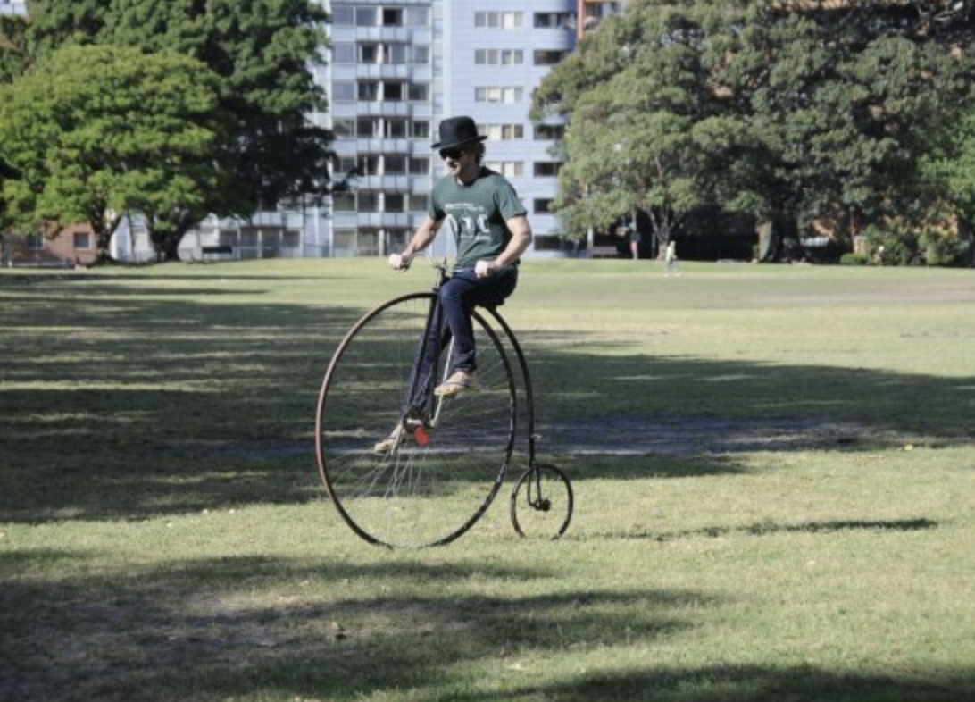 Person riding a vintage penny-farthing bicycle in a park with trees and buildings in the background.