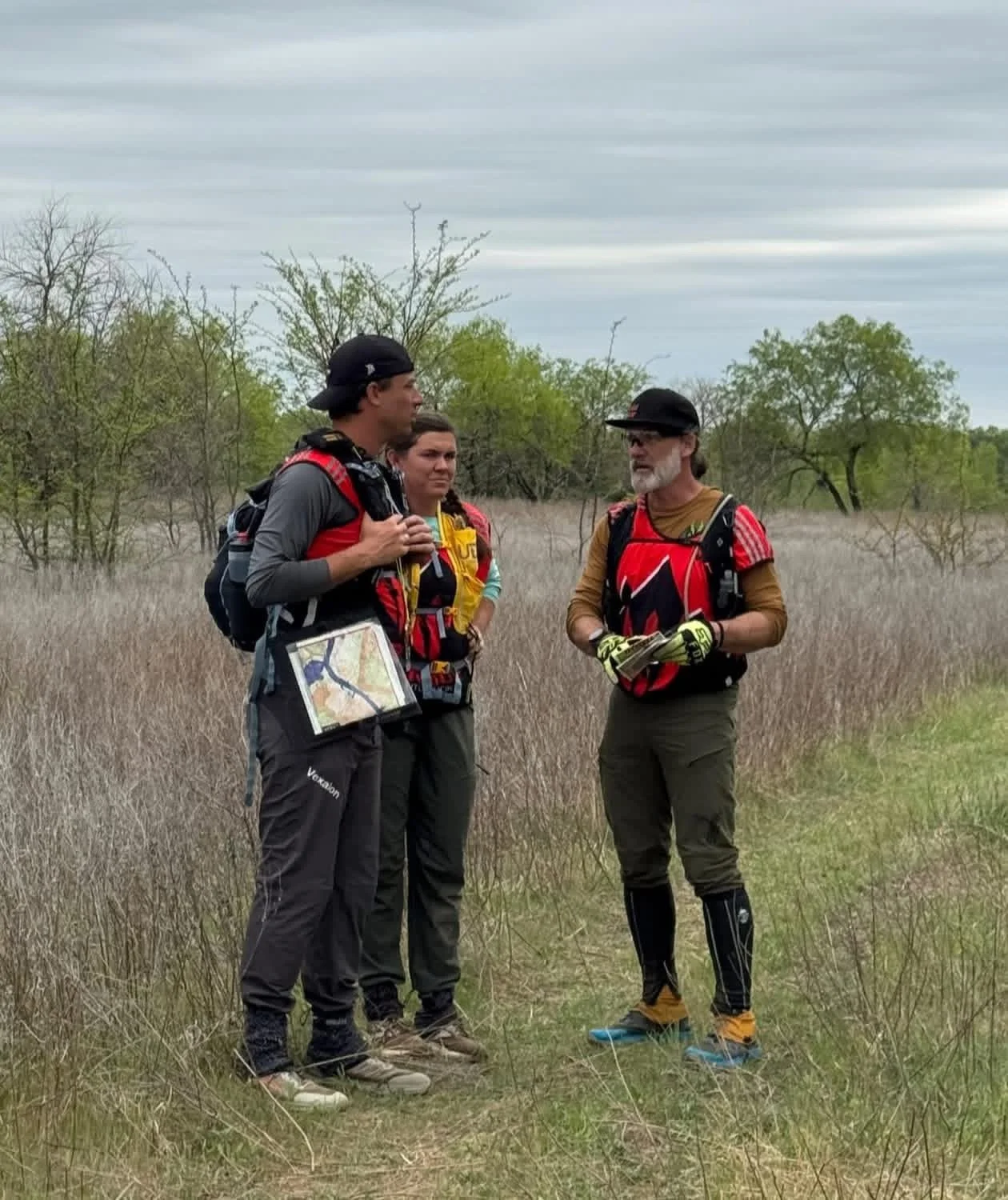 Four Adventure Racers dressed in outdoor gear and backpacks standing in a field of tall grass during a race, having a conversation during daytime with overcast sky, with trees in the background.