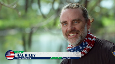 A smiling man with a beard and long hair tied back, wearing a bandana with American flag colors, standing outdoors with blurred greenery in the background, possibly on a boat or near water.