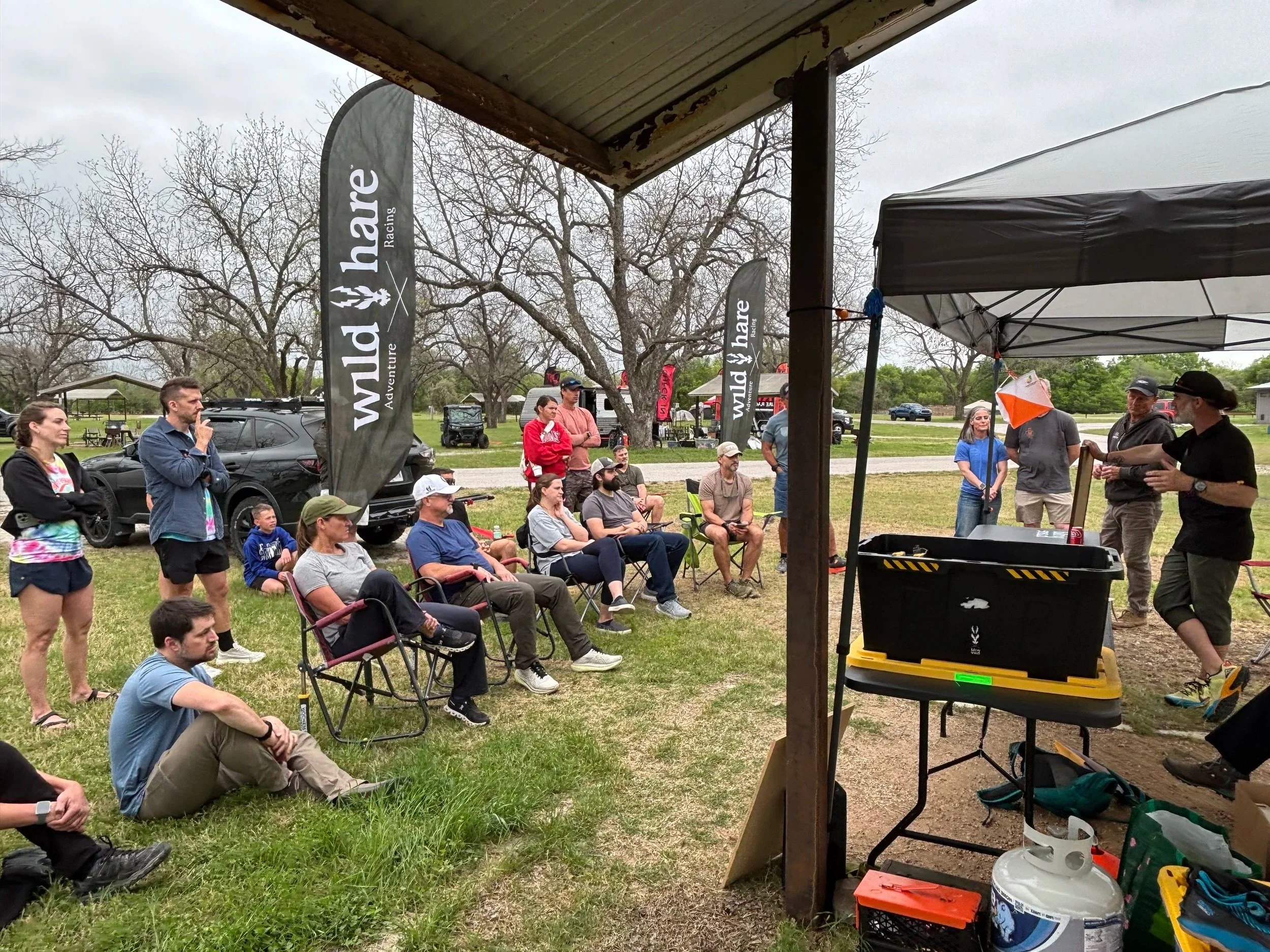 People gathered outdoors at a racetrack event, sitting on chairs and standing under a canopy, with trees and parked vehicles in the background.