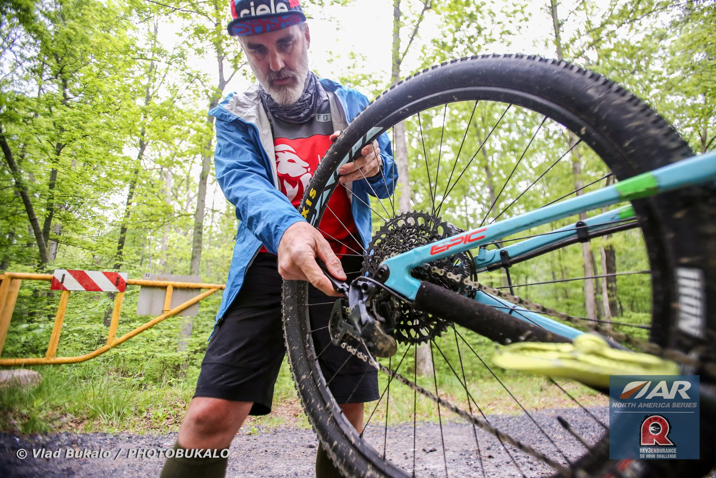 Hal Riley, captain of the Adventure Racing team is fixing  a mountain bike in a wooded outdoor area with green leaves. During the USARA North America Series.