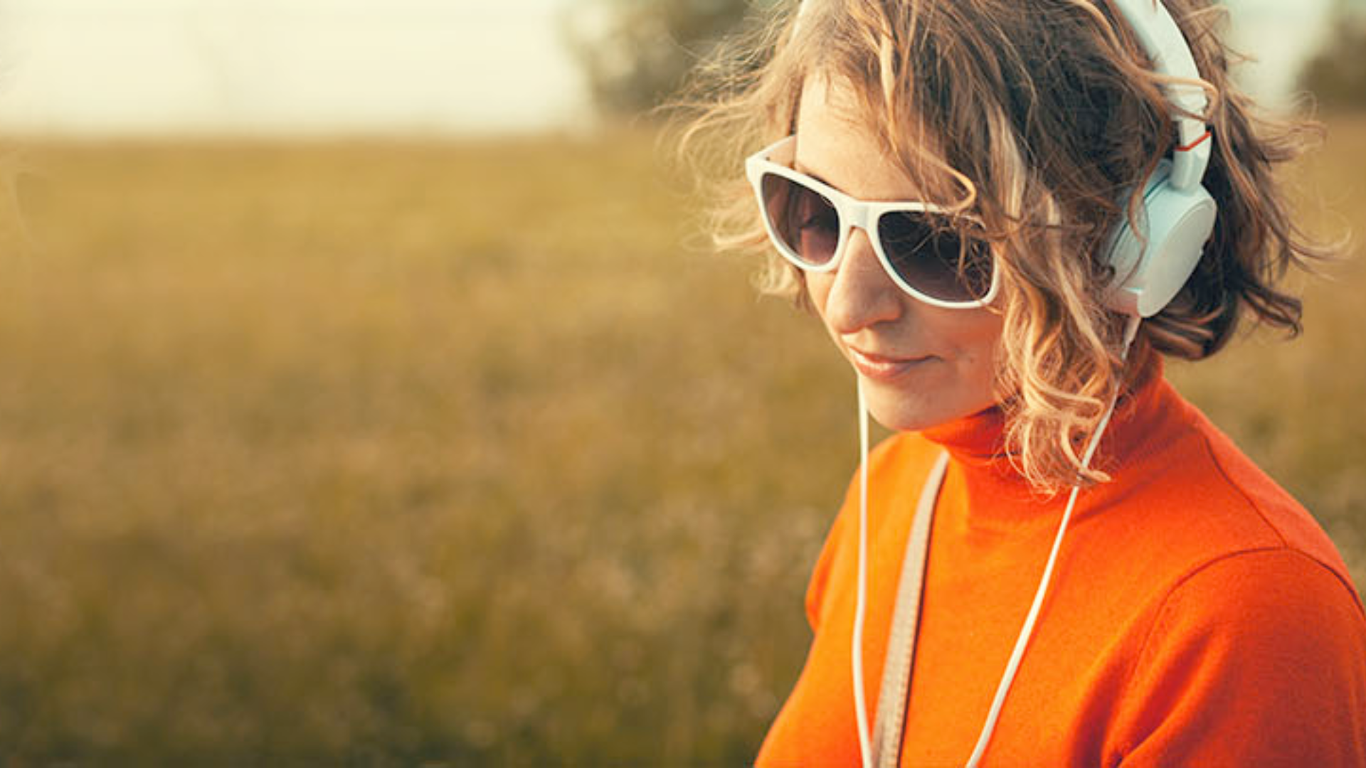 Woman in sunglasses and headphones wearing orange top in grassy field