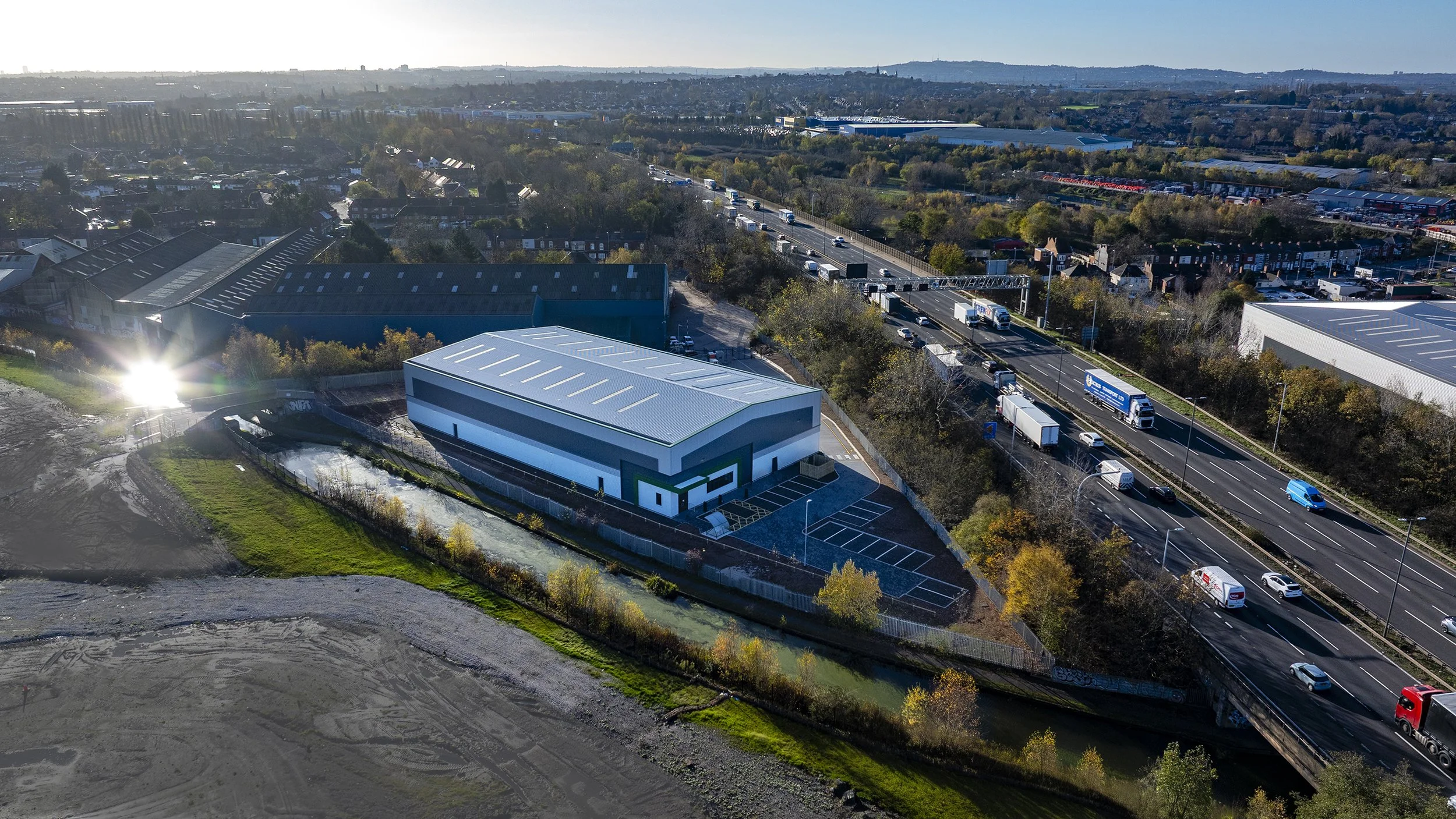 Aerial view of a modern building with parking lot, next to a busy highway with cars and trucks, surrounded by trees and industrial areas.
