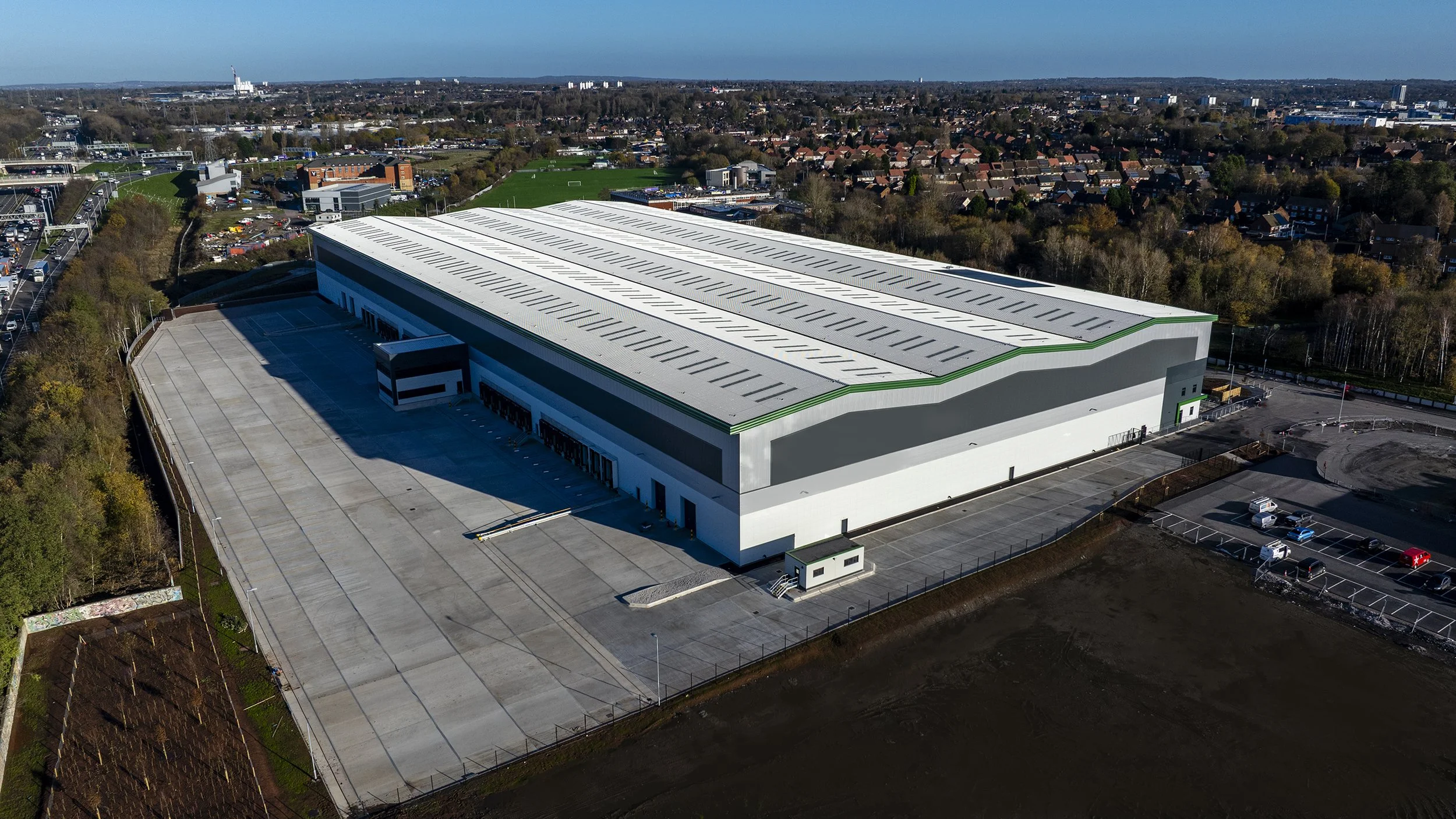 Aerial view of a large modern warehouse with a white and gray exterior, green accents, and a vast parking lot surrounded by trees and a cityscape in the background.
