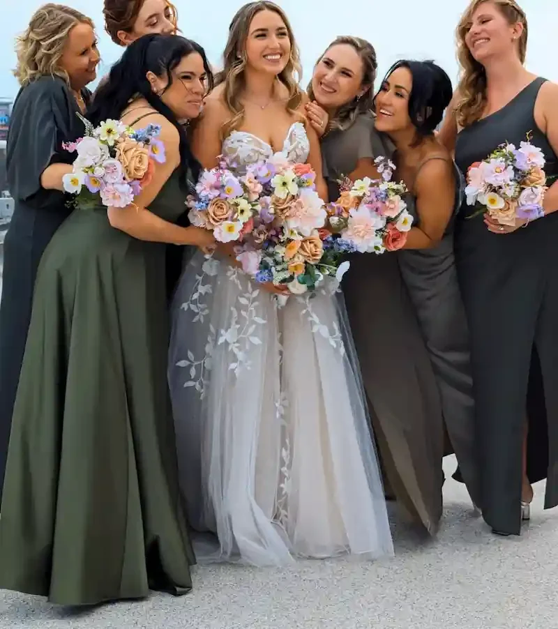 Bride surrounded by bridesmaids in olive and sage gowns holding colorful silk floral bouquets featuring peach roses, coral dahlias, lavender blooms, and wildflowers from the Penelope collection — Floréal Blooms NJ