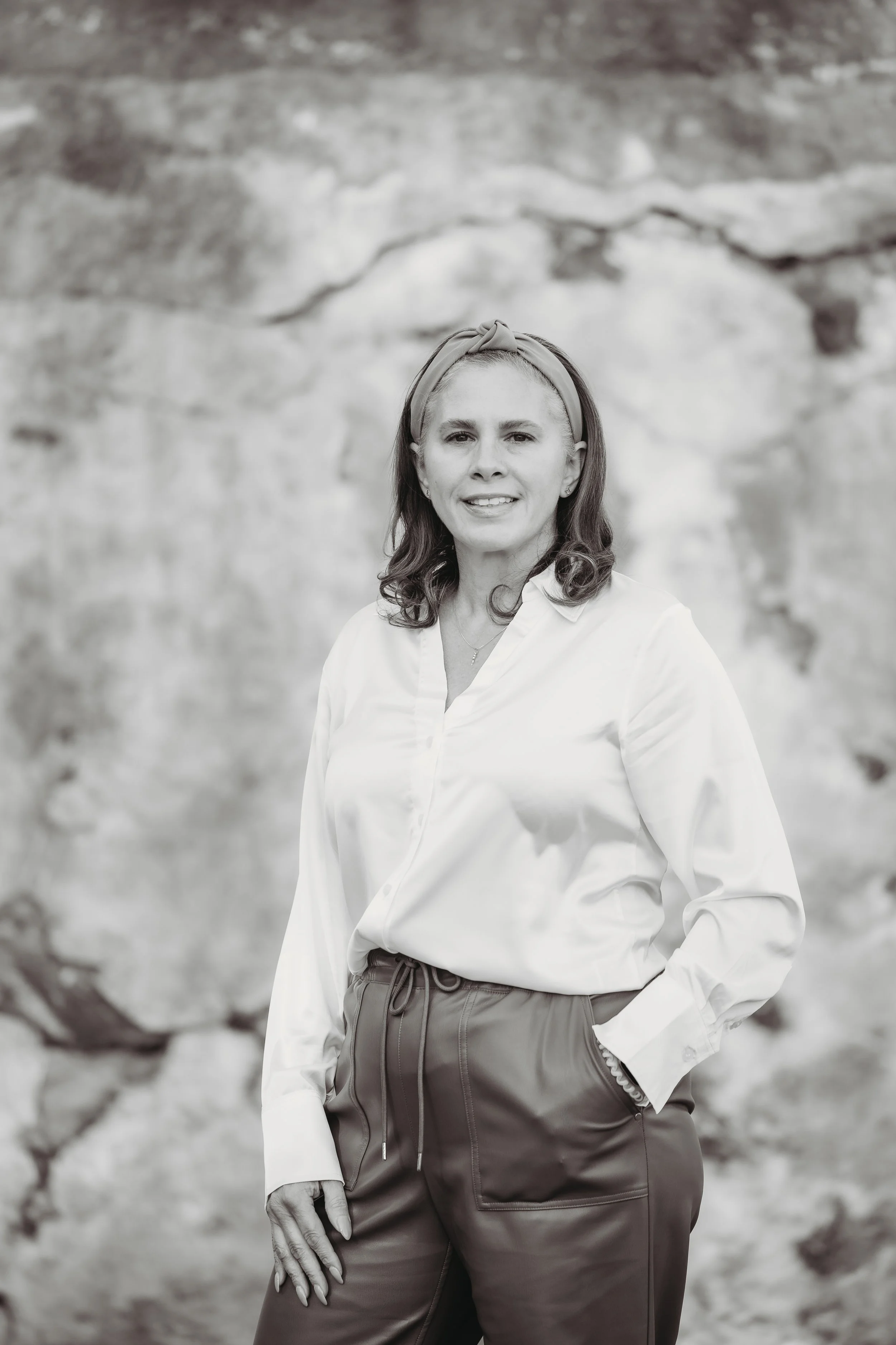 Black and white photograph of Melissa Fife - owner & CEO of events by mwith shoulder-length hair, wearing a headband, a white button-up shirt, and pants, standing in front of a rocky background. She has one hand in her pocket and is smiling slightly.