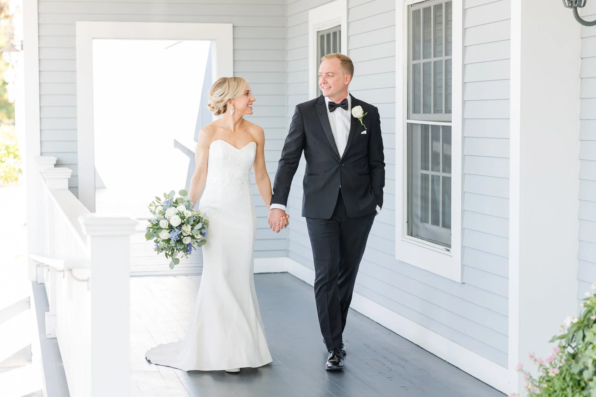 Bride and groom walking hand in hand outside a house, bride holding a bouquet of white and purple flowers, bride in a strapless white wedding dress, groom in a black tuxedo with a bow tie and white boutonniere.