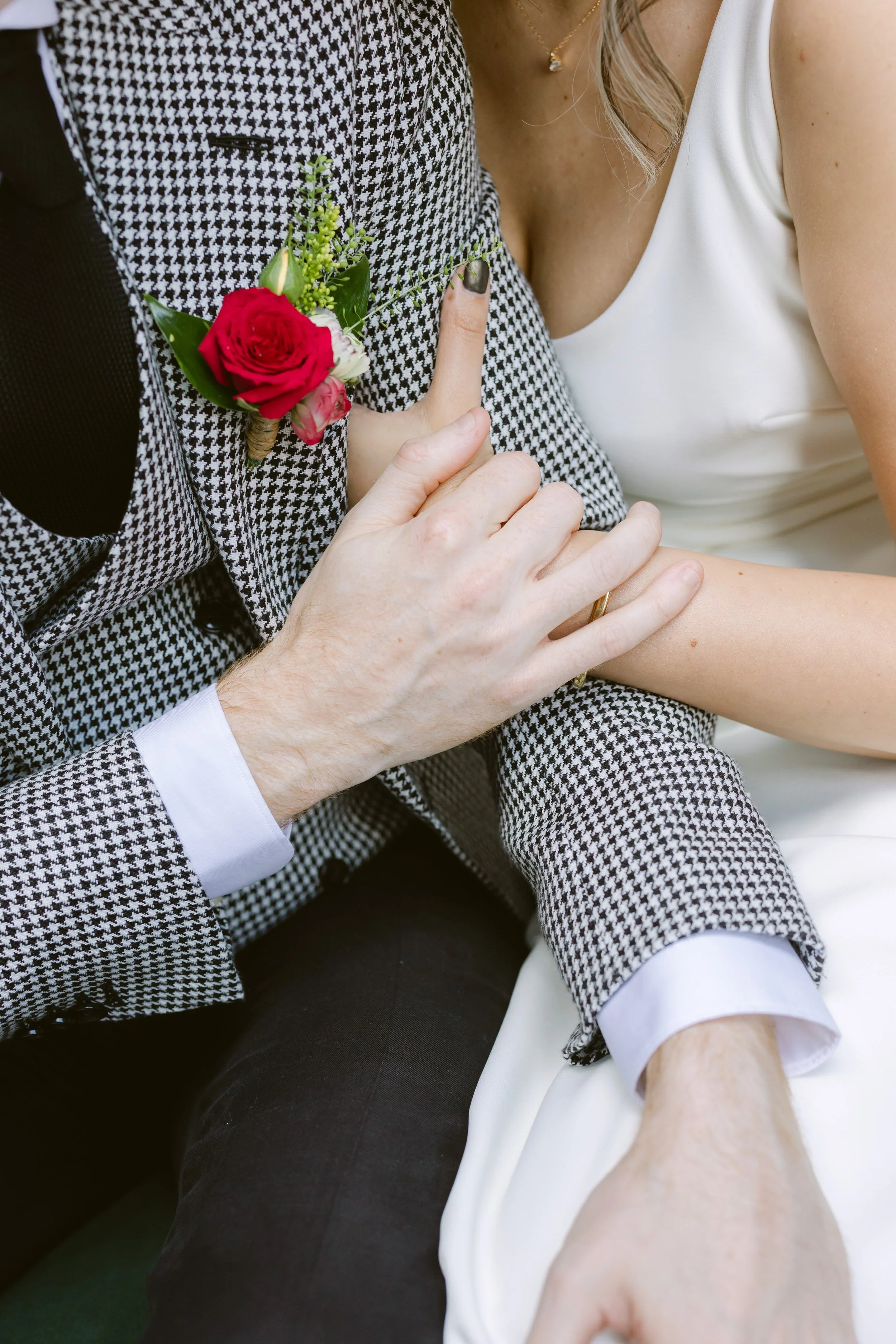 Close-up of a groom in a black-and-white checked suit with a red rose boutonniere, gently holding the hand of a bride dressed in white, during a wedding ceremony or celebration.