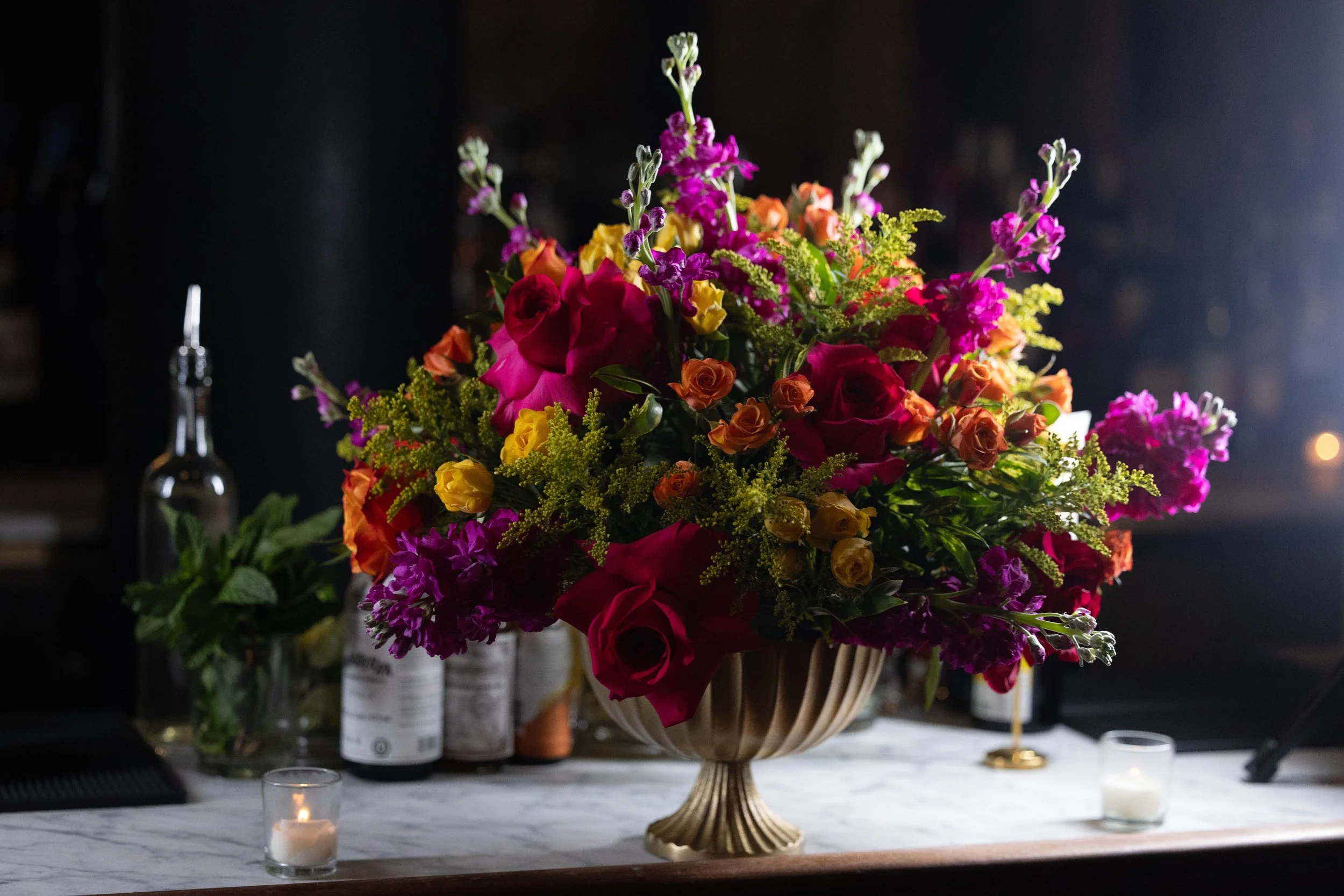 A large colorful bouquet of flowers in a decorative vase on a white marble countertop, with small candles and bottles in the background.