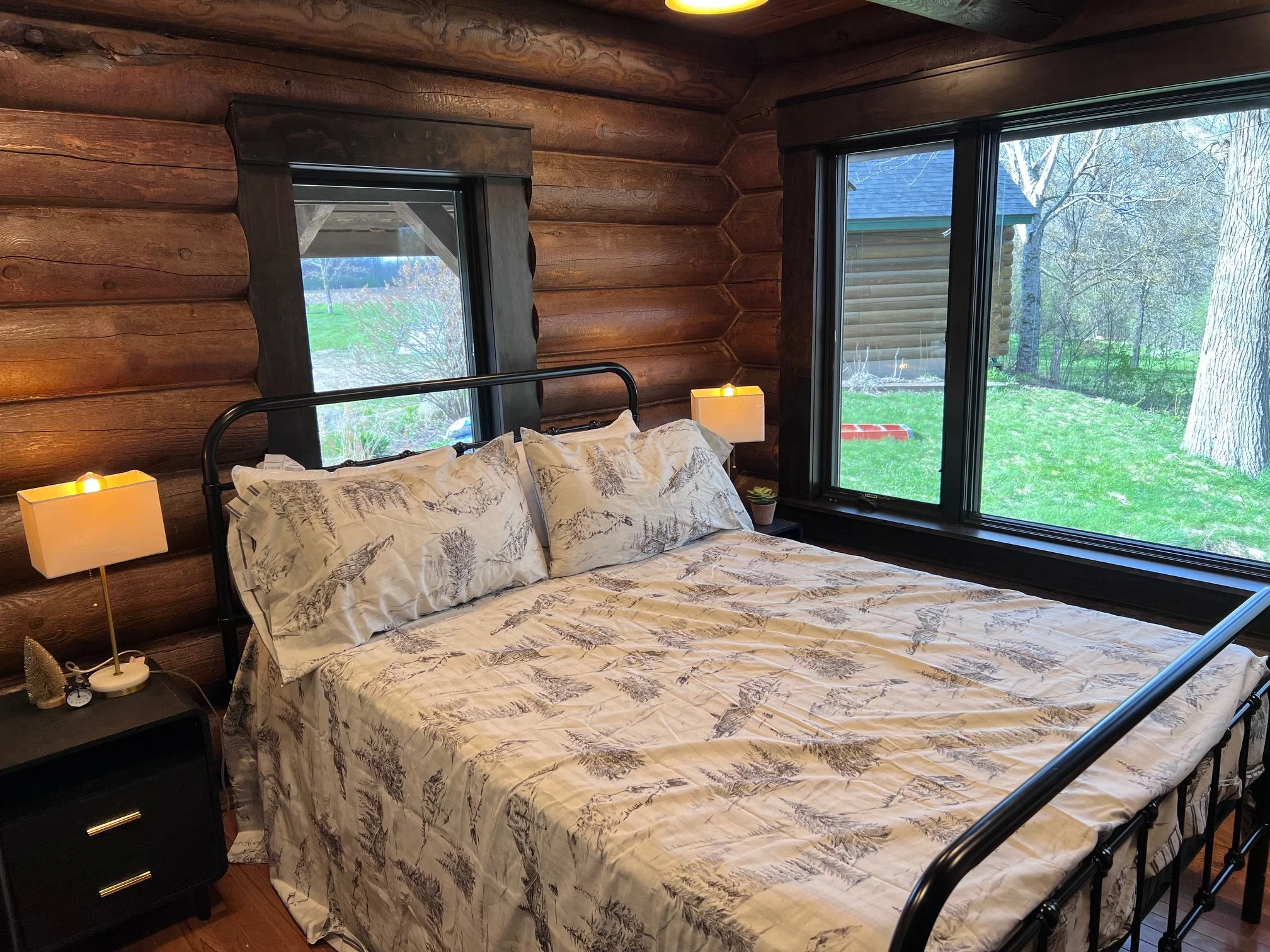 A cozy rustic bedroom with a bed covered in nature-themed sheets and pillows, dark wood nightstand with a lamp, large windows showing a green outdoor scene with trees and grass, and wooden log walls.