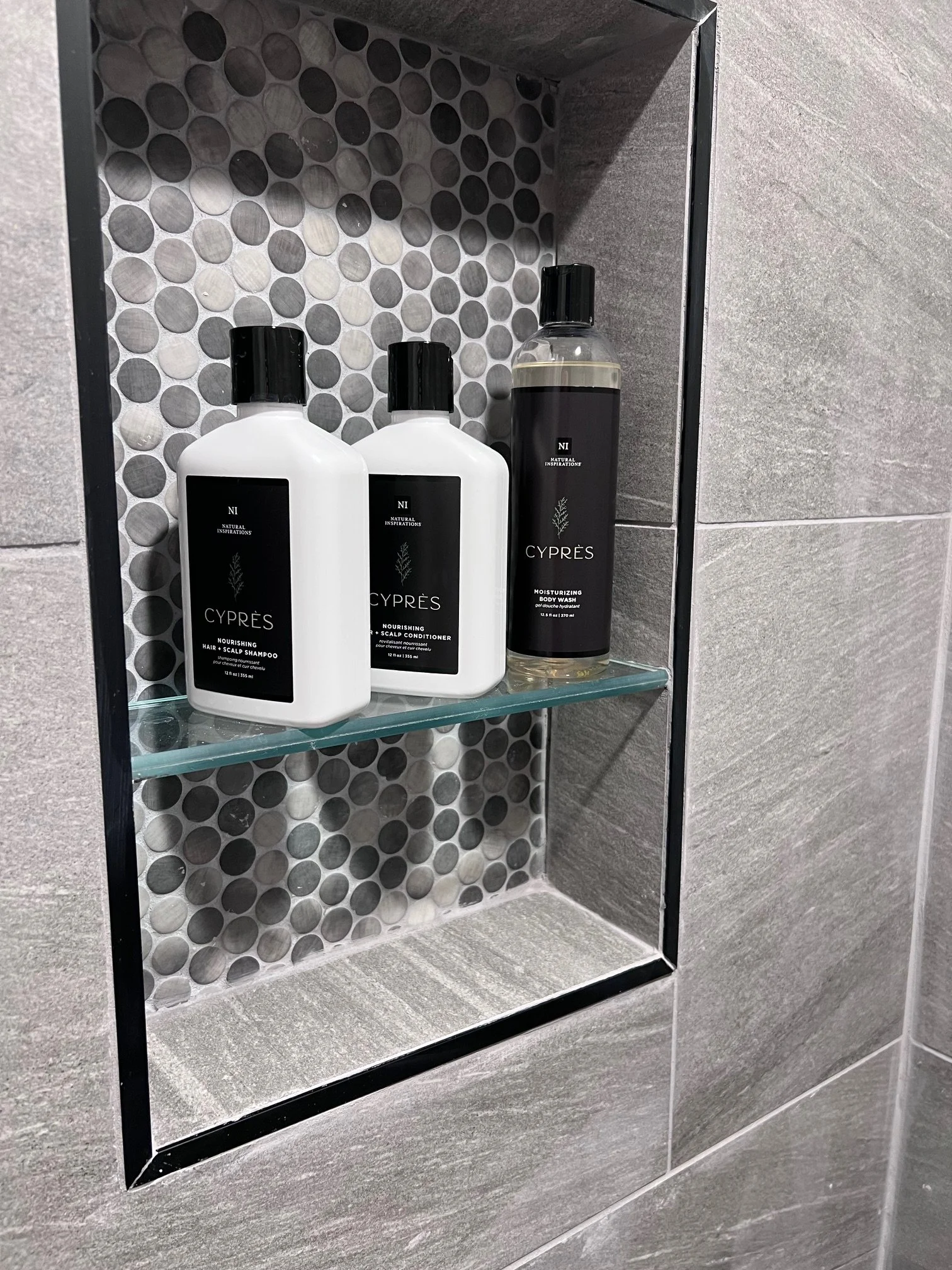 A shower niche with three bottles of hair and body products on a glass shelf, surrounded by gray tiled walls with a circular mosaic pattern inside.