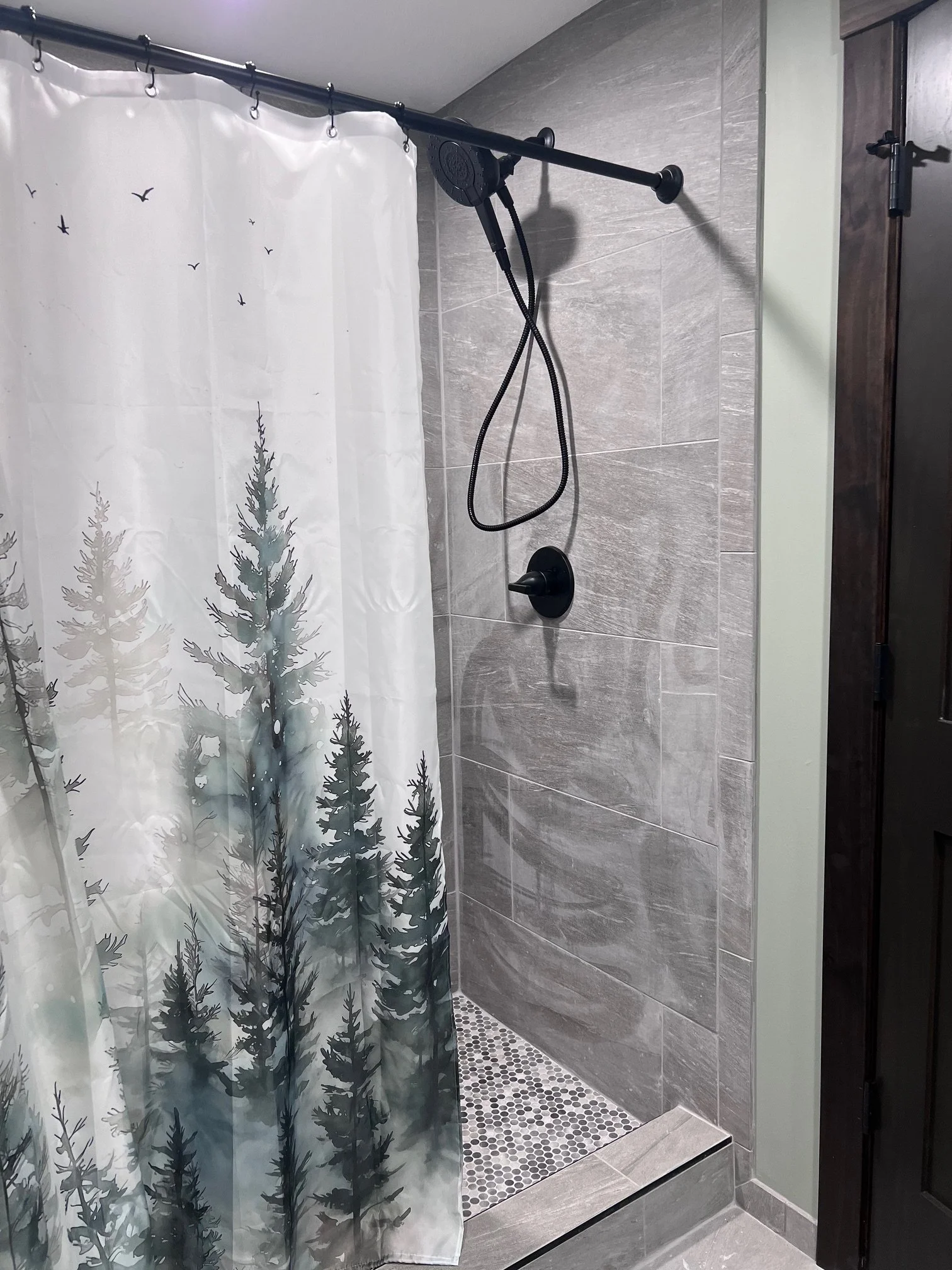 Shower area with gray textured tile walls, a black shower head and hose, a black shower fixture, a white shower curtain with pine trees and flying birds design, and small hexagonal black and white floor tiles.