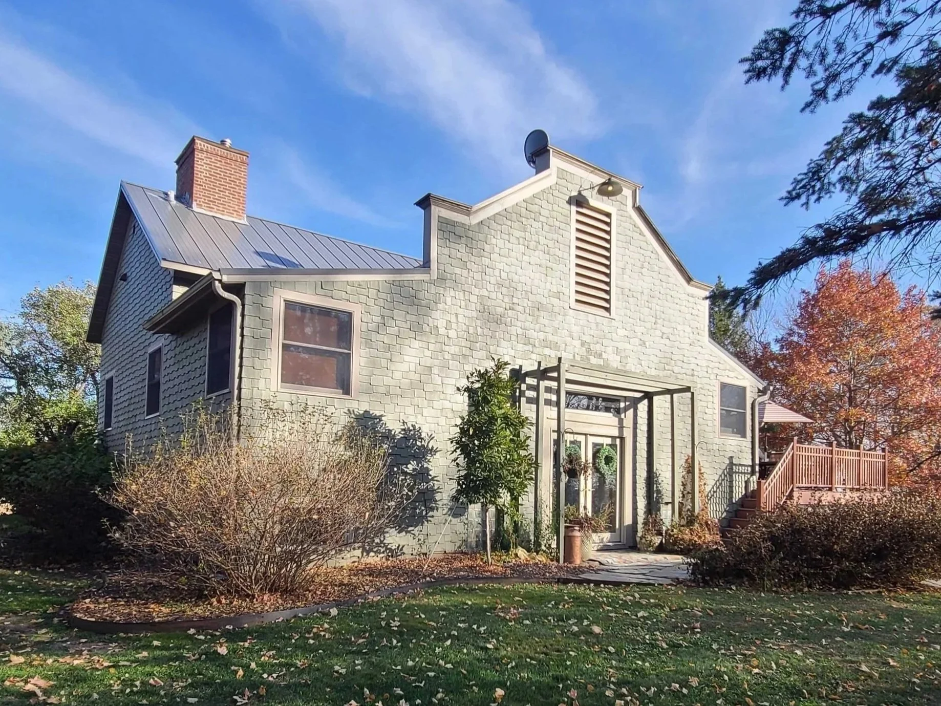 A two-story house with grey shingle siding, brick chimney, metal roof, and a small porch with a wreath on the door, surrounded by a garden with trees showing fall colors and a clear blue sky in the background.
