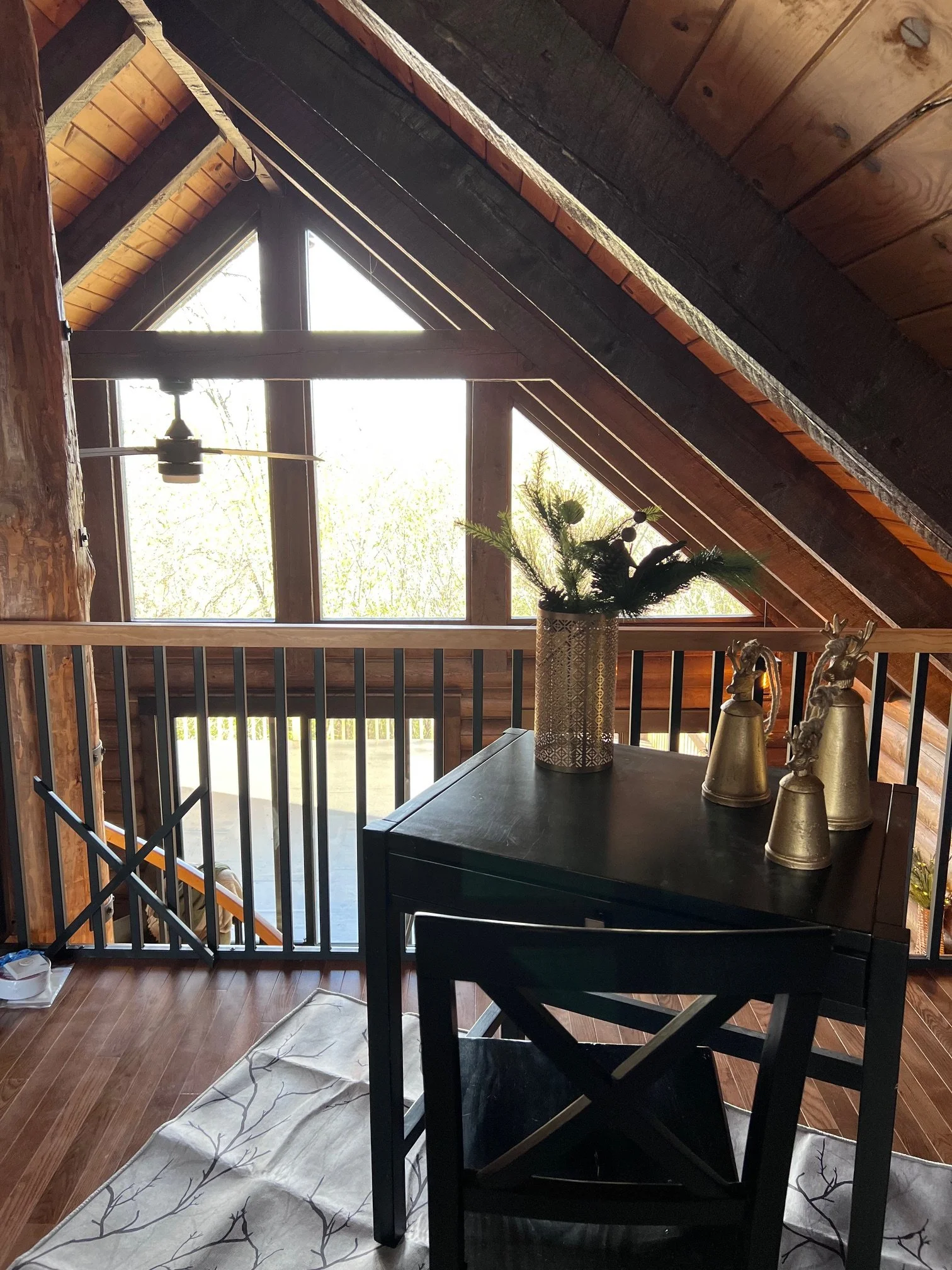 A cozy attic room with large triangular windows letting in natural light. It features a dark wooden table with decorative items including gold bells and a vase with greenery. The ceiling has exposed wooden beams, and the walls are lined with wood pan