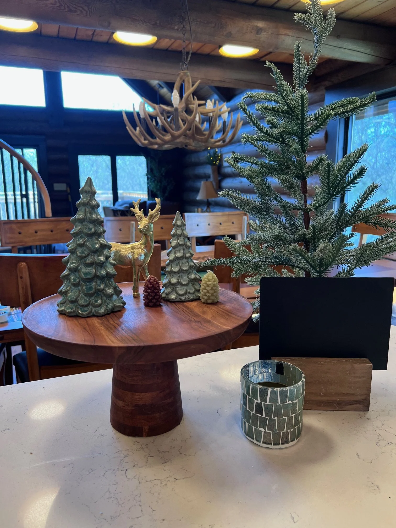 Decorative holiday table centerpiece with ceramic trees, a gold reindeer figurine, a pine cone, and a cylindrical mosaic-patterned candle holder. In the background, a small Christmas tree and festive decor are visible in a cozy wooden interior.
