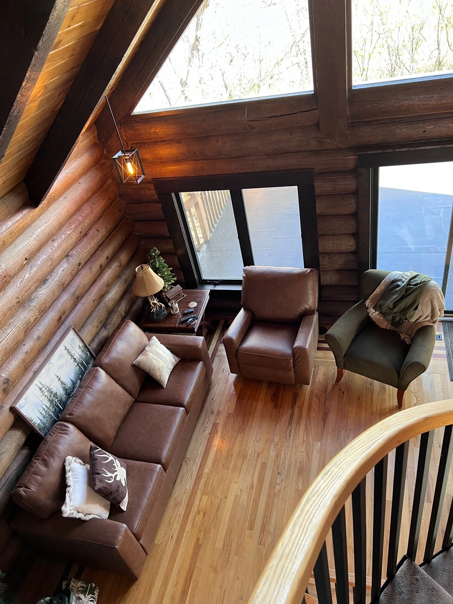 Interior view of a cozy log cabin living room with wooden walls and ceiling, large window, leather sofa with decorative pillows, a wooden side table with a lamp and decoration, and a green armchair with a blanket. The ceiling has a hanging light fixt