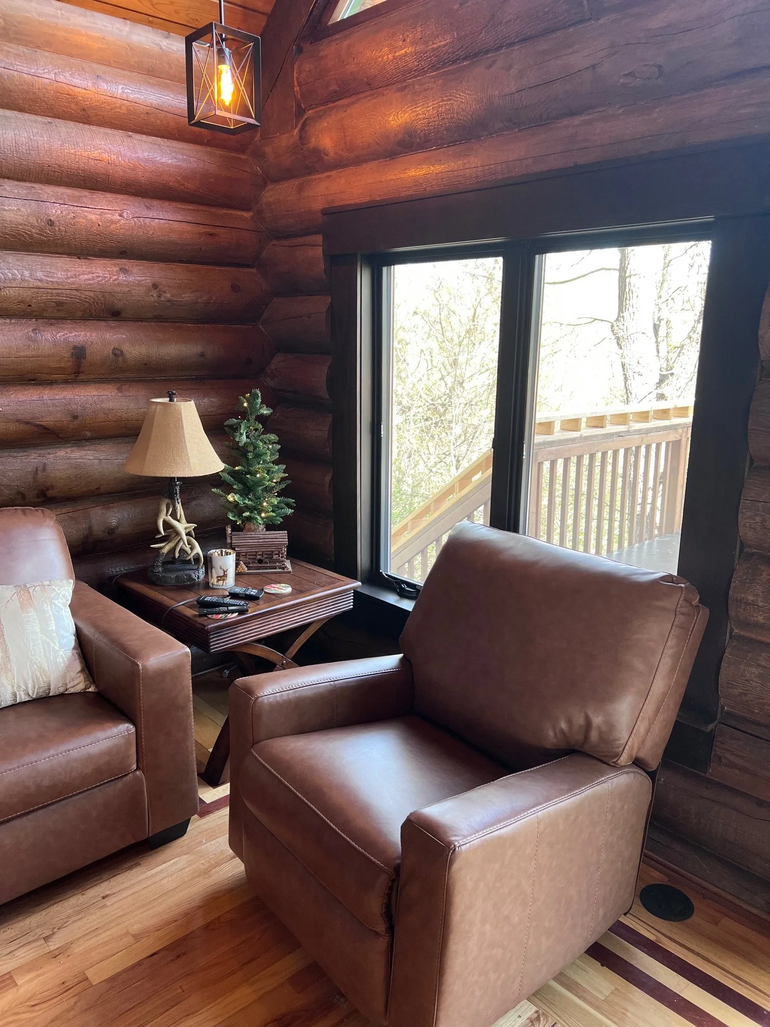 A cozy living room with leather armchairs, a wooden coffee table, and a small Christmas tree with decorations. There is a large window leading to a balcony with trees outside. The walls are made of wooden logs, and a geometric pendant light hangs fro